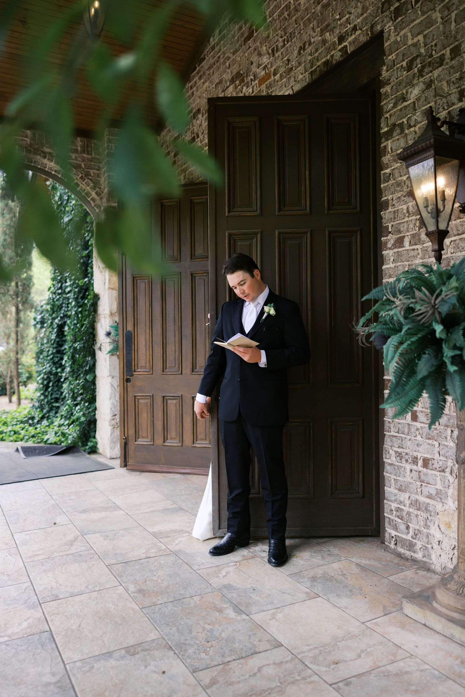 Groom in black tuxedo reading vows outside elegant brick venue with ivy-covered walls and ornate wooden doors.