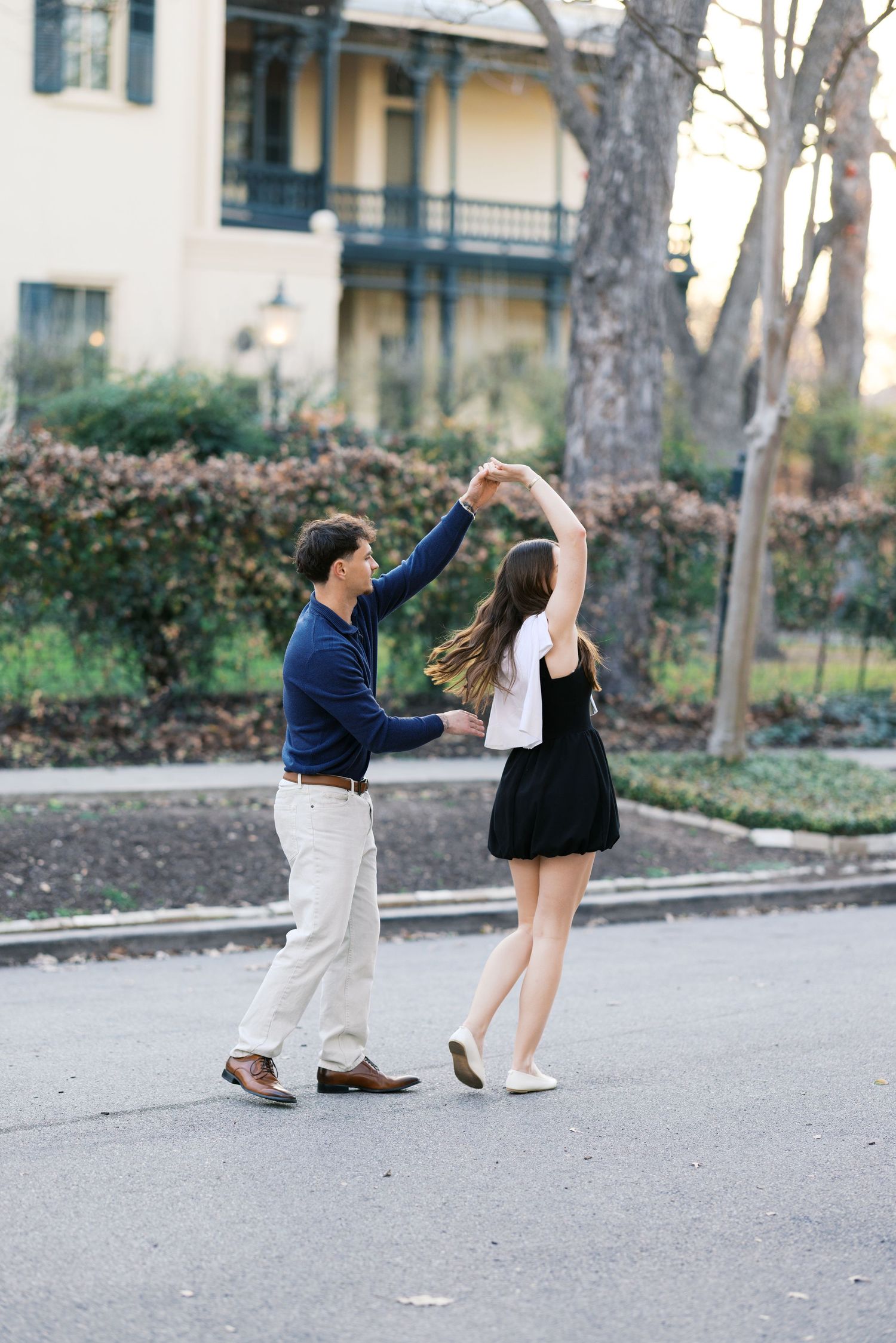 Couple dancing outdoors on a street, man spinning woman in black skirt and white top near a historic building.