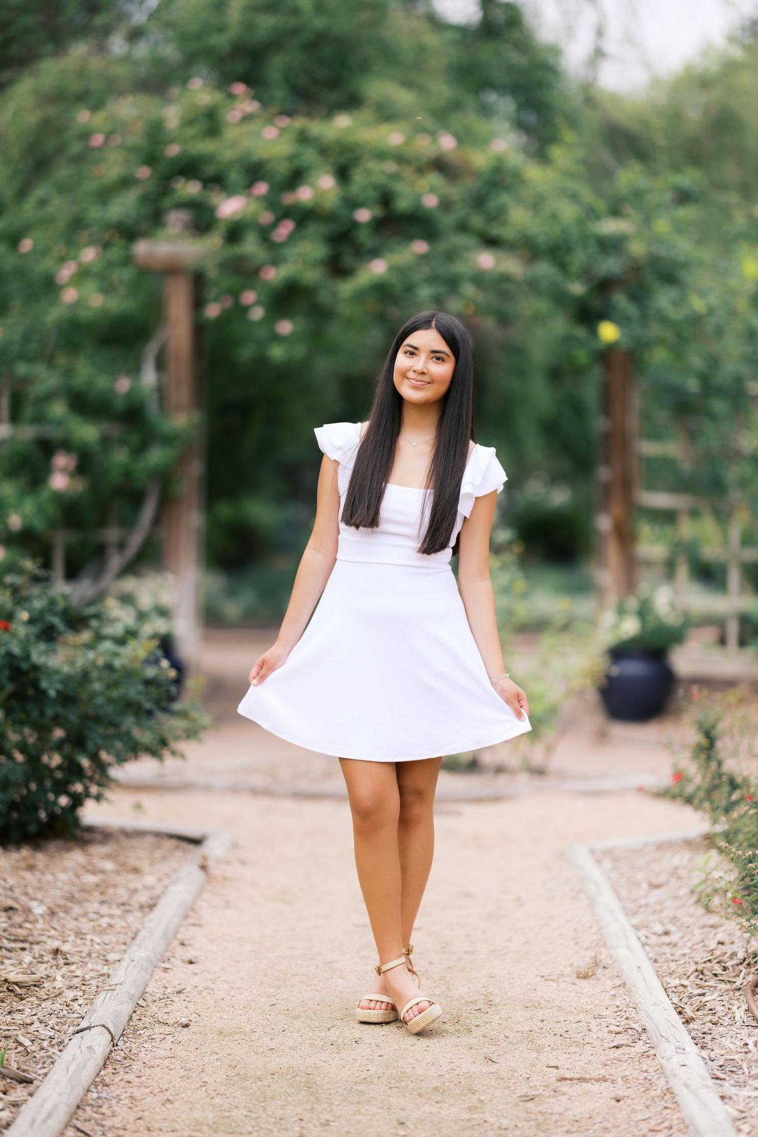 Young woman in white dress walking on garden path surrounded by lush greenery and roses.