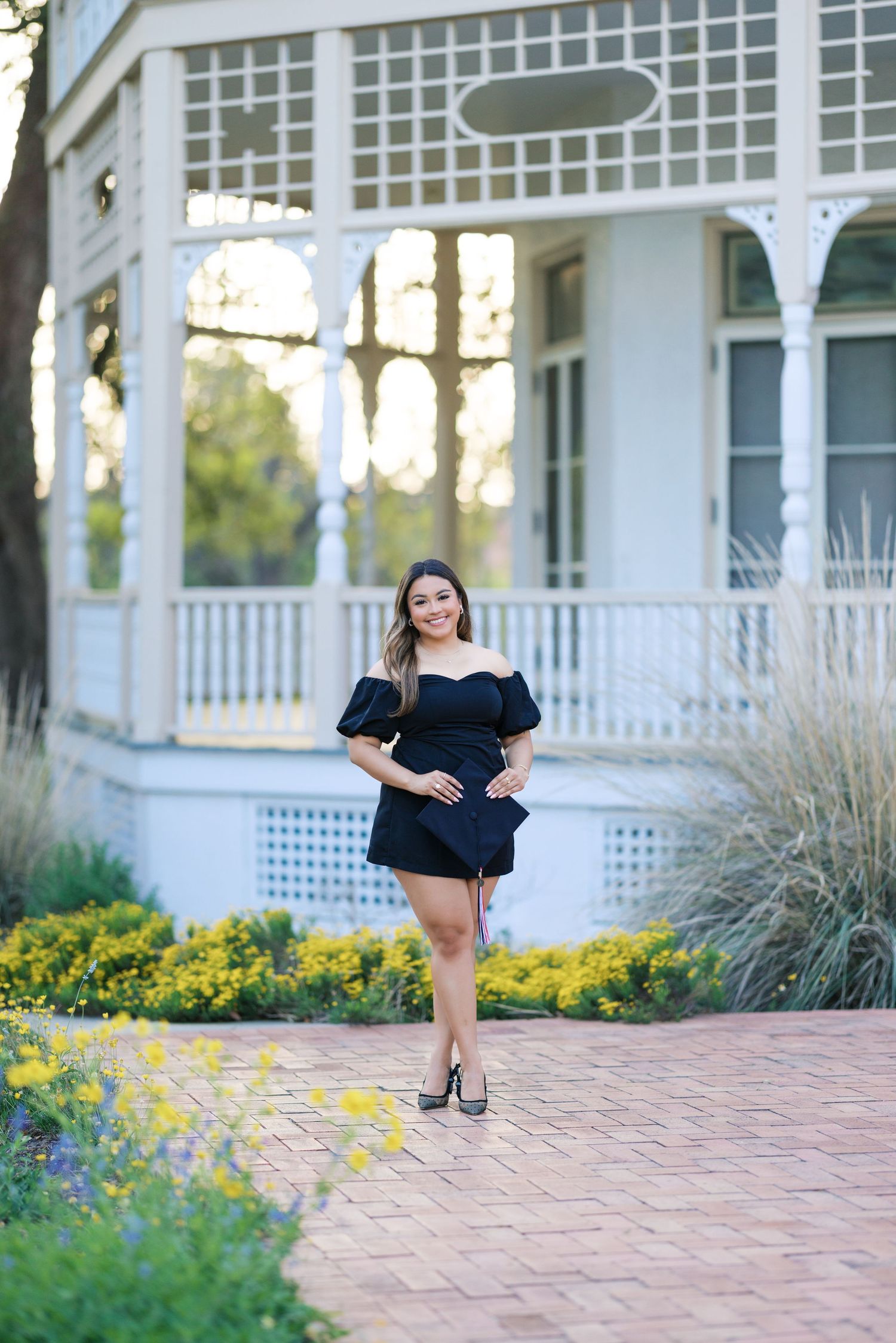 Young woman in black off-shoulder mini dress posing on brick path by white Victorian gazebo with yellow flowers.