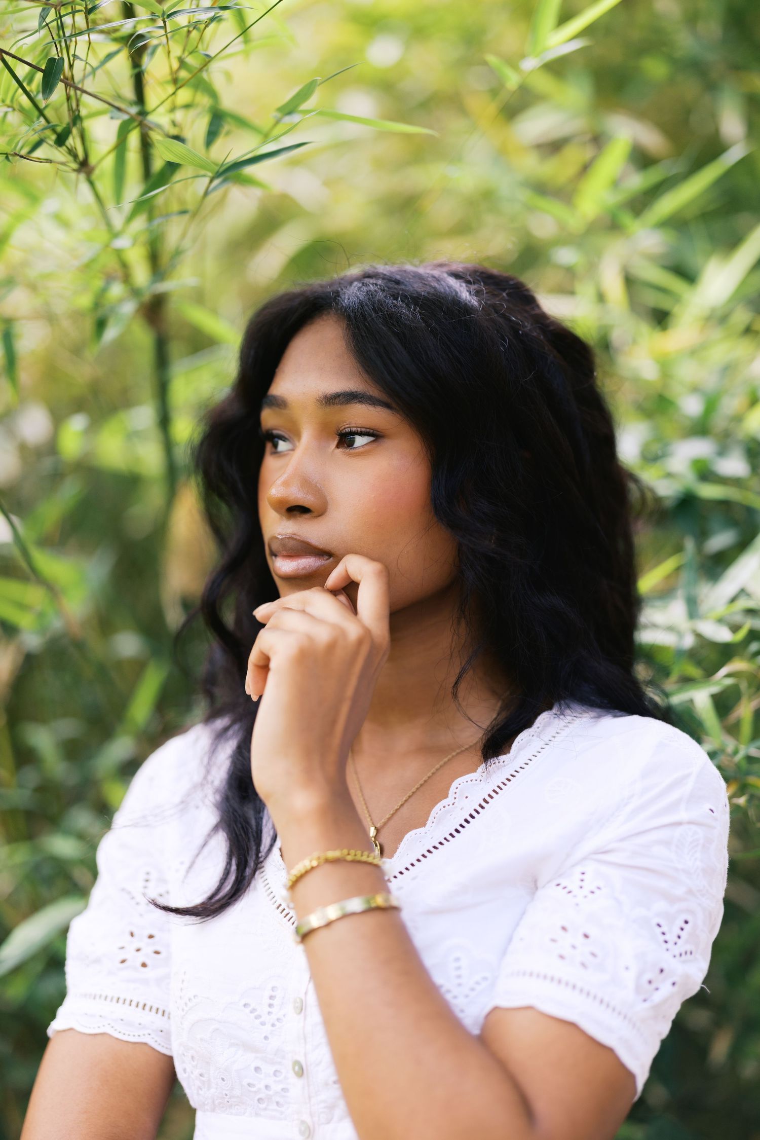 Young Black woman in white eyelet top gazing thoughtfully amid green bamboo leaves.