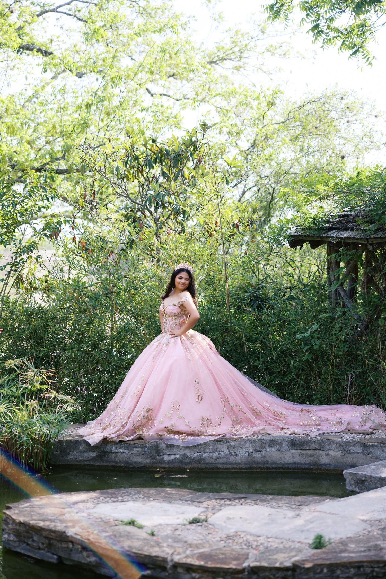 Young woman in a pink ball gown quinceañera dress poses gracefully in a lush garden setting.