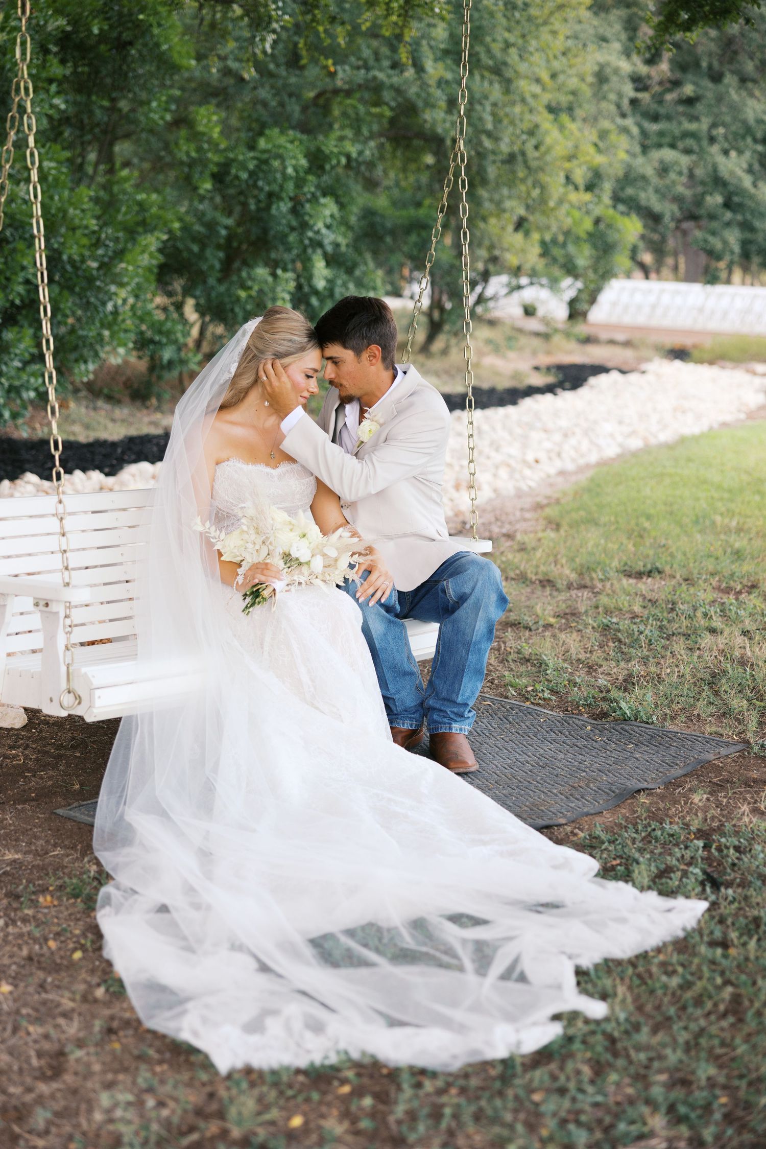 Romantic couple on white wooden swing with flowing wedding dress in garden setting.