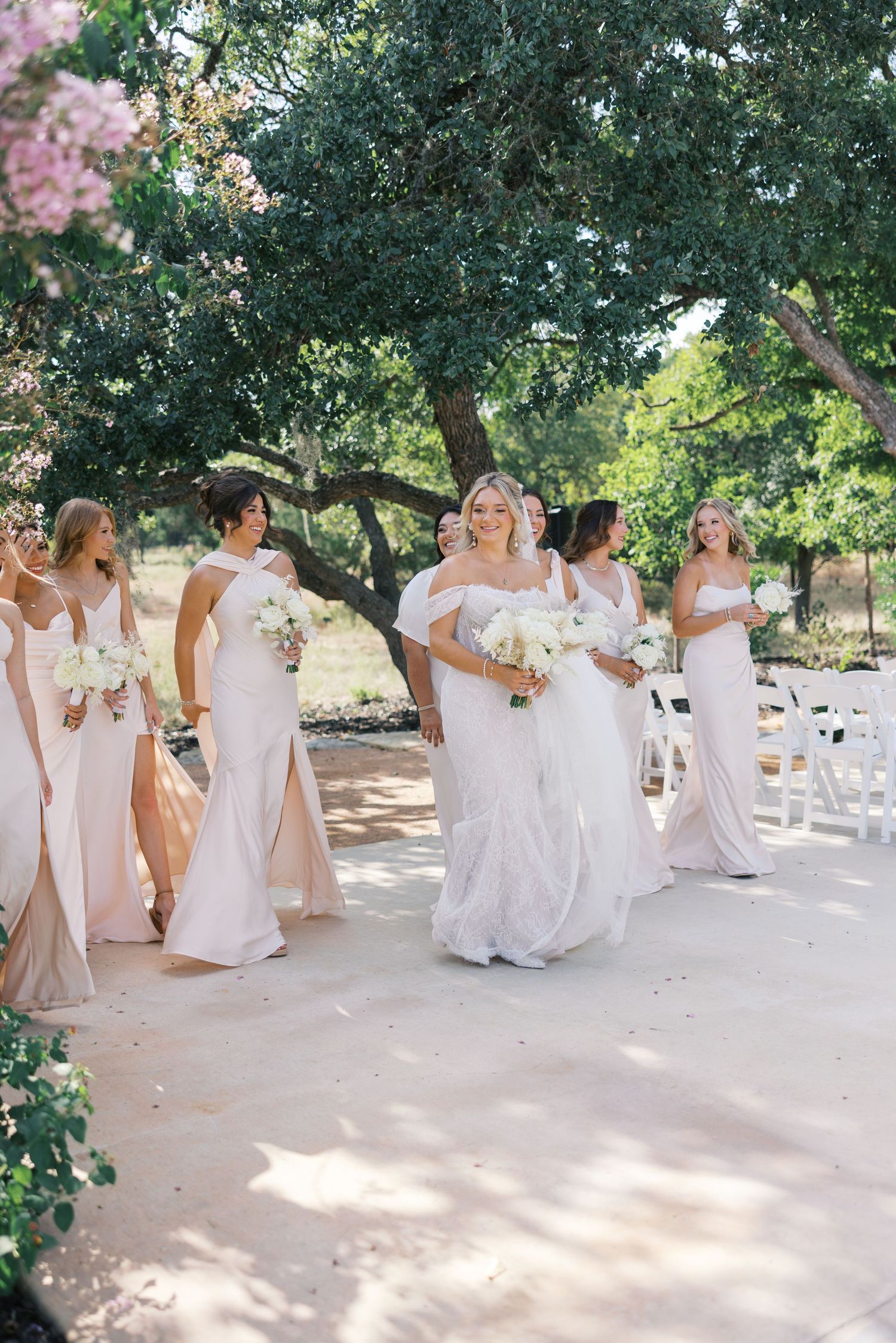 Bridesmaids in flowing white and blush dresses walking together outdoors under trees.