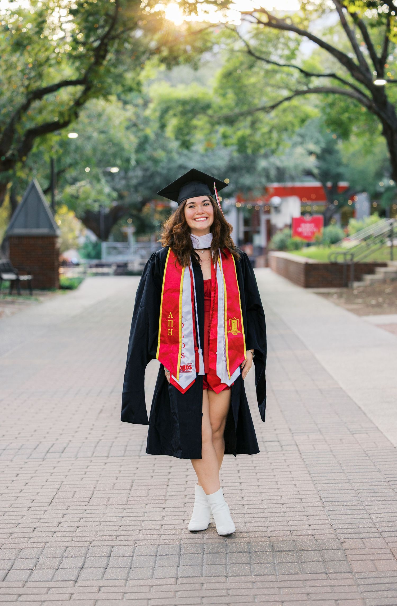 Graduate in cap and gown with red stoles walking on brick pathway surrounded by trees.