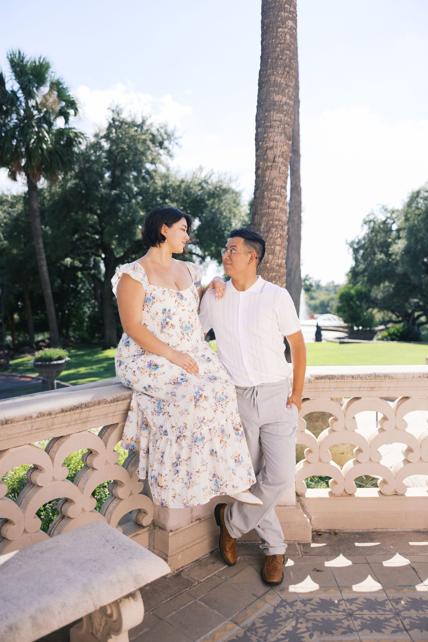 A couple sits together on a stone balcony surrounded by palm trees on a sunny day.