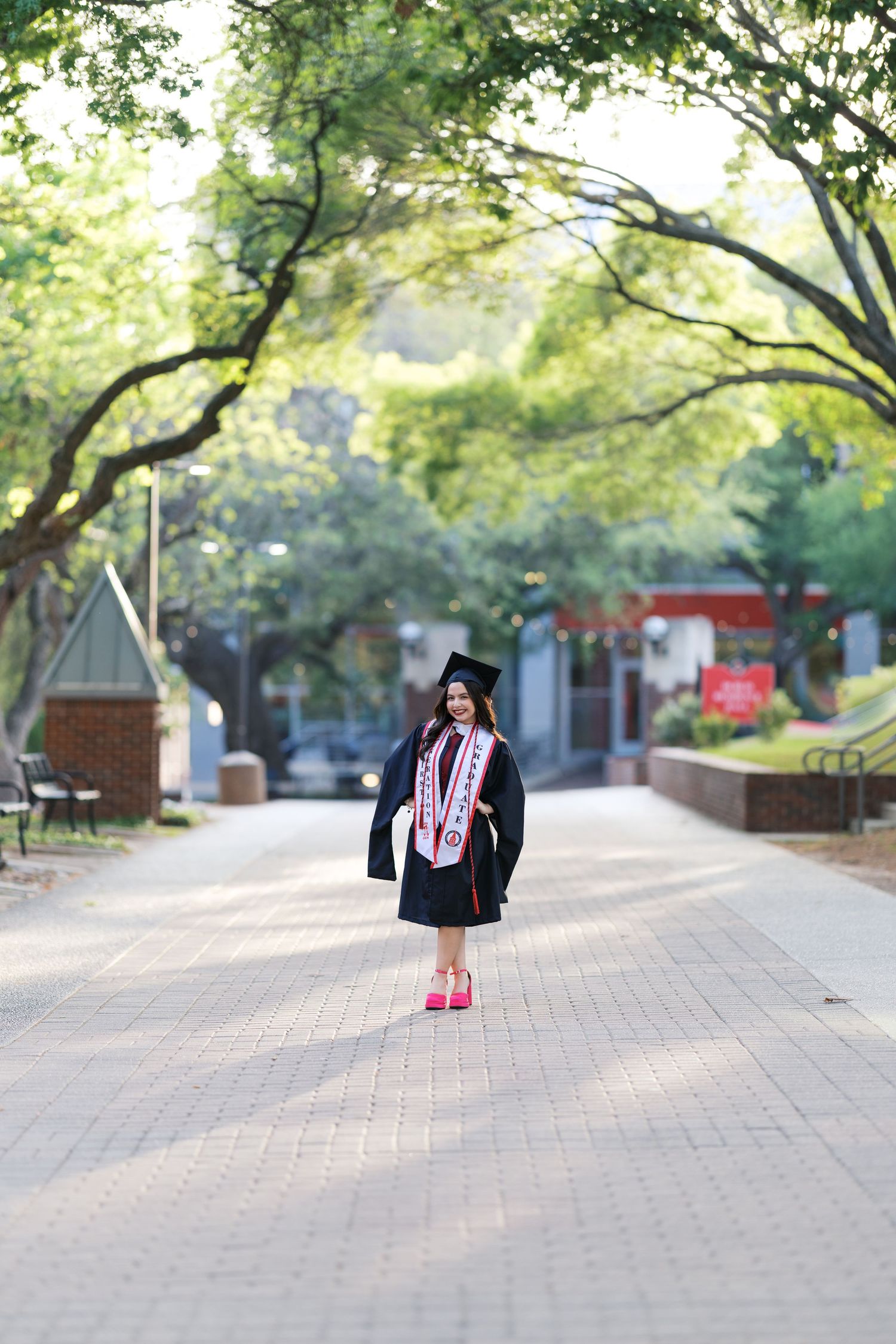Smiling graduate in black cap and gown tosses stole on tree-lined brick university campus path.