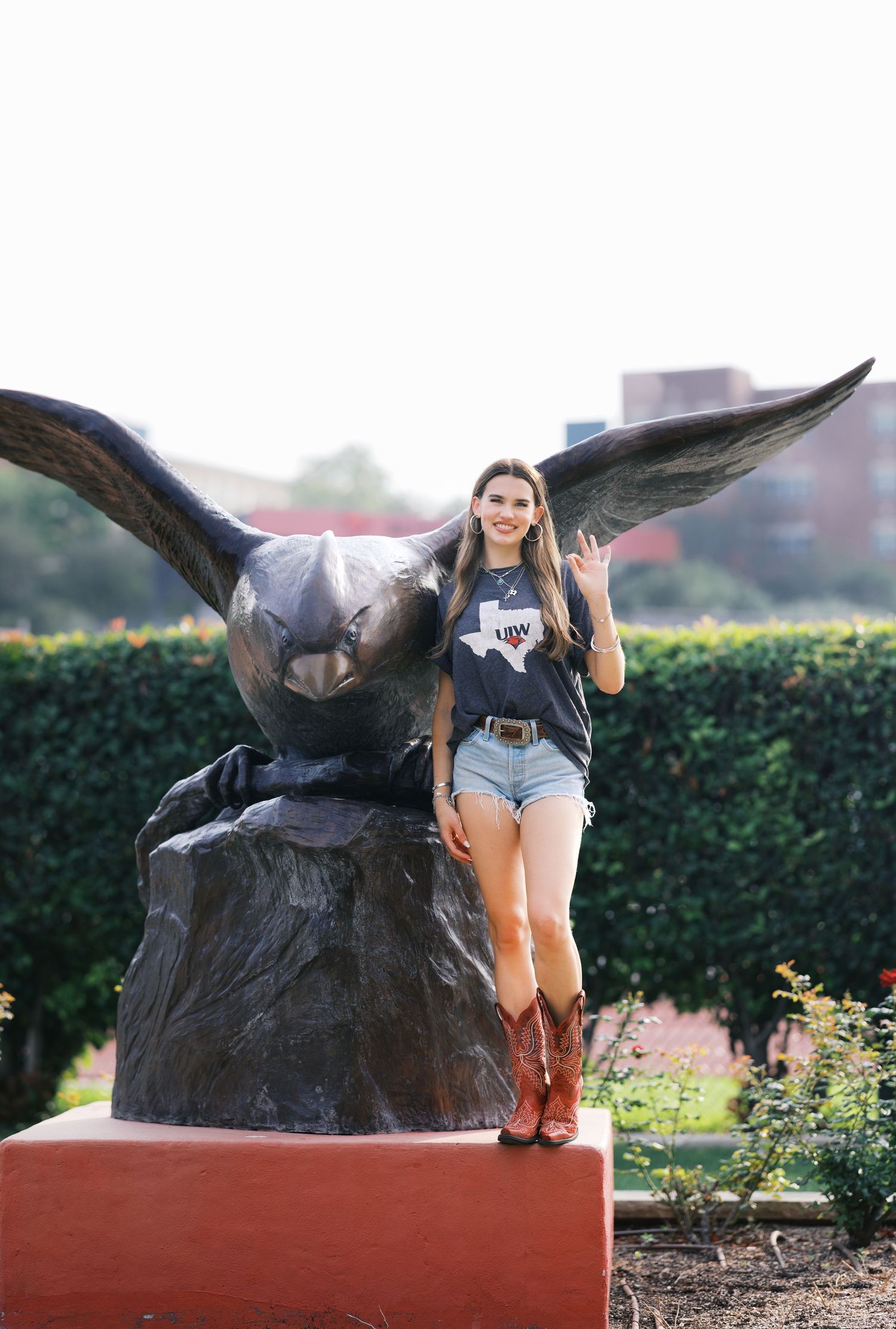Young woman in cowboy boots and denim shorts posing beside a large bronze eagle statue outdoors.