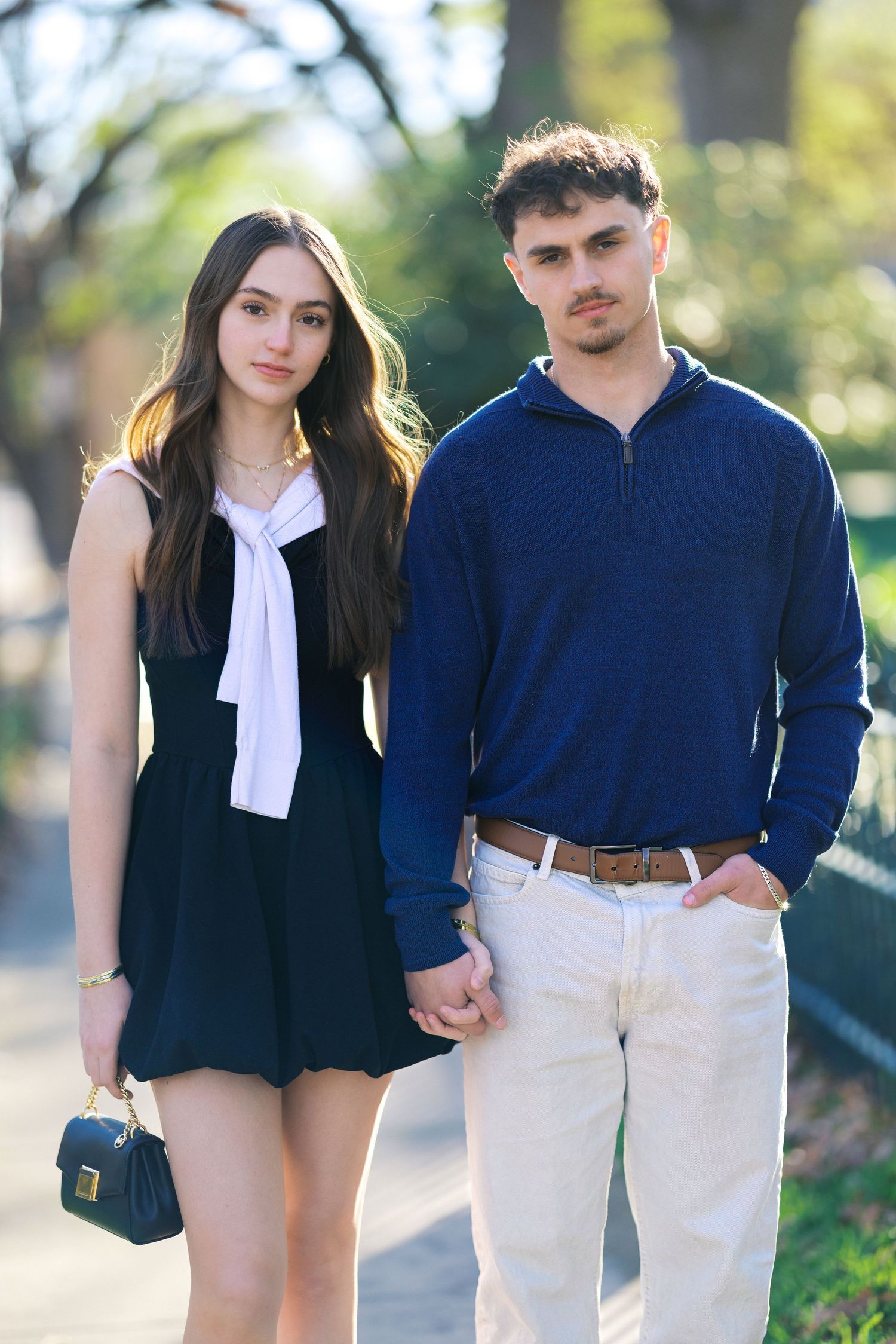 Stylish young couple holding hands on a park path, woman in black mini dress with white bow tie.