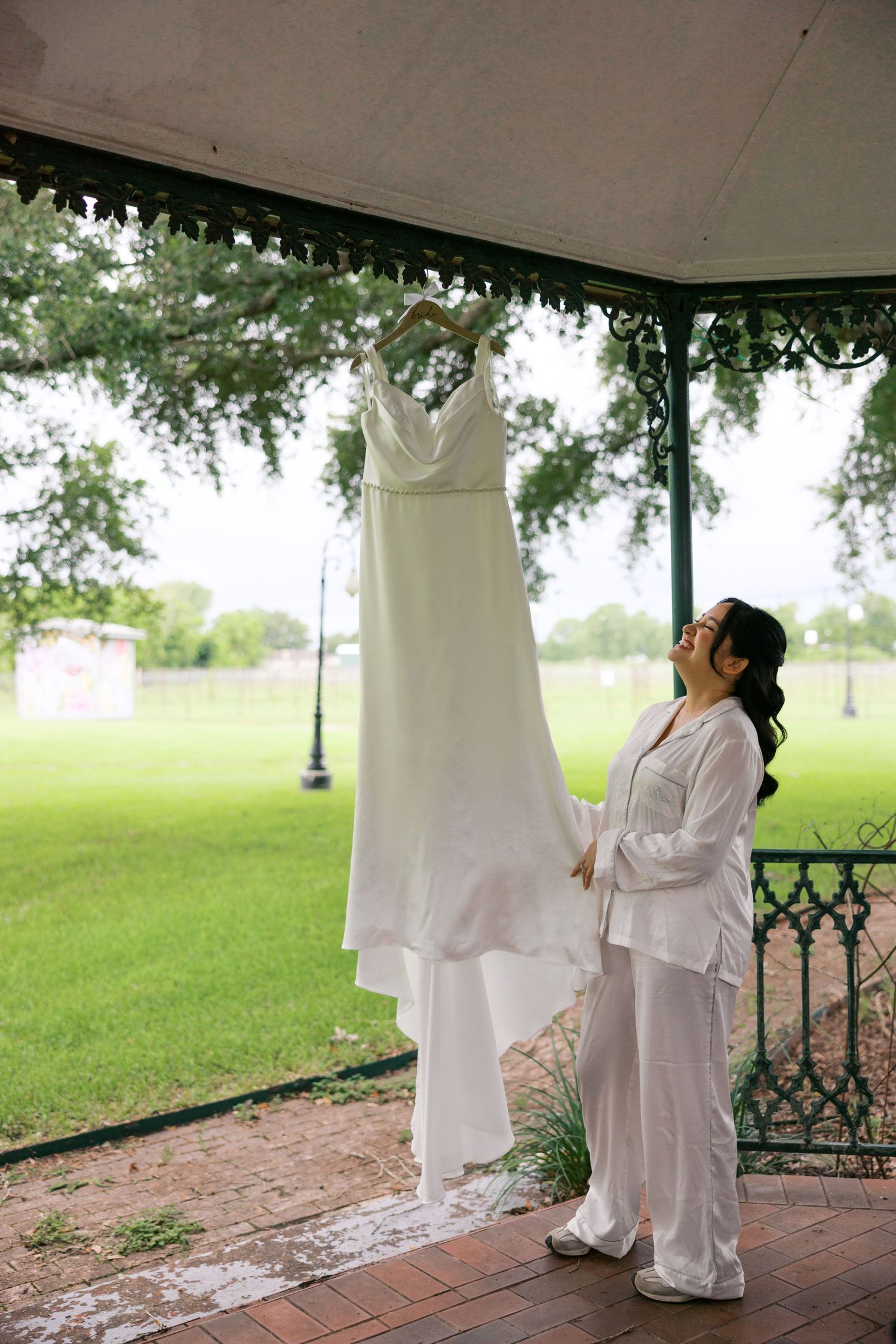 Bride in white pajamas admiring her wedding dress hanging in a gazebo surrounded by lush greenery.