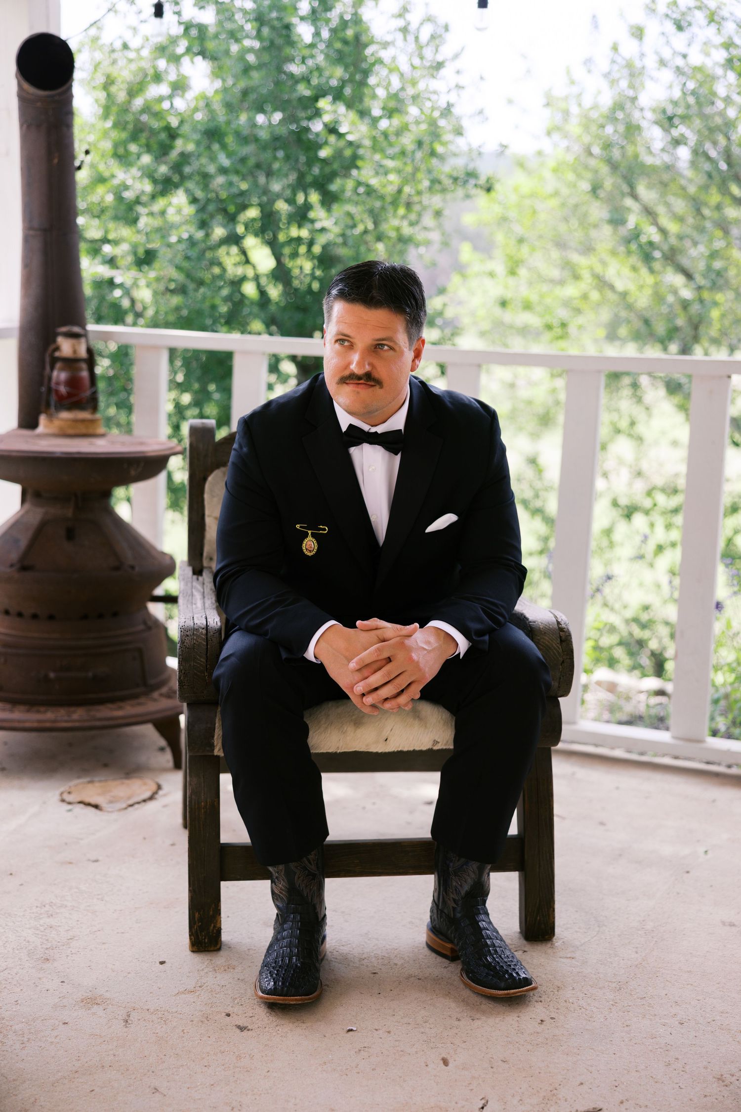 Groom in black tuxedo with bow tie sits in wooden chair on porch, hands clasped, with lush green trees behind.