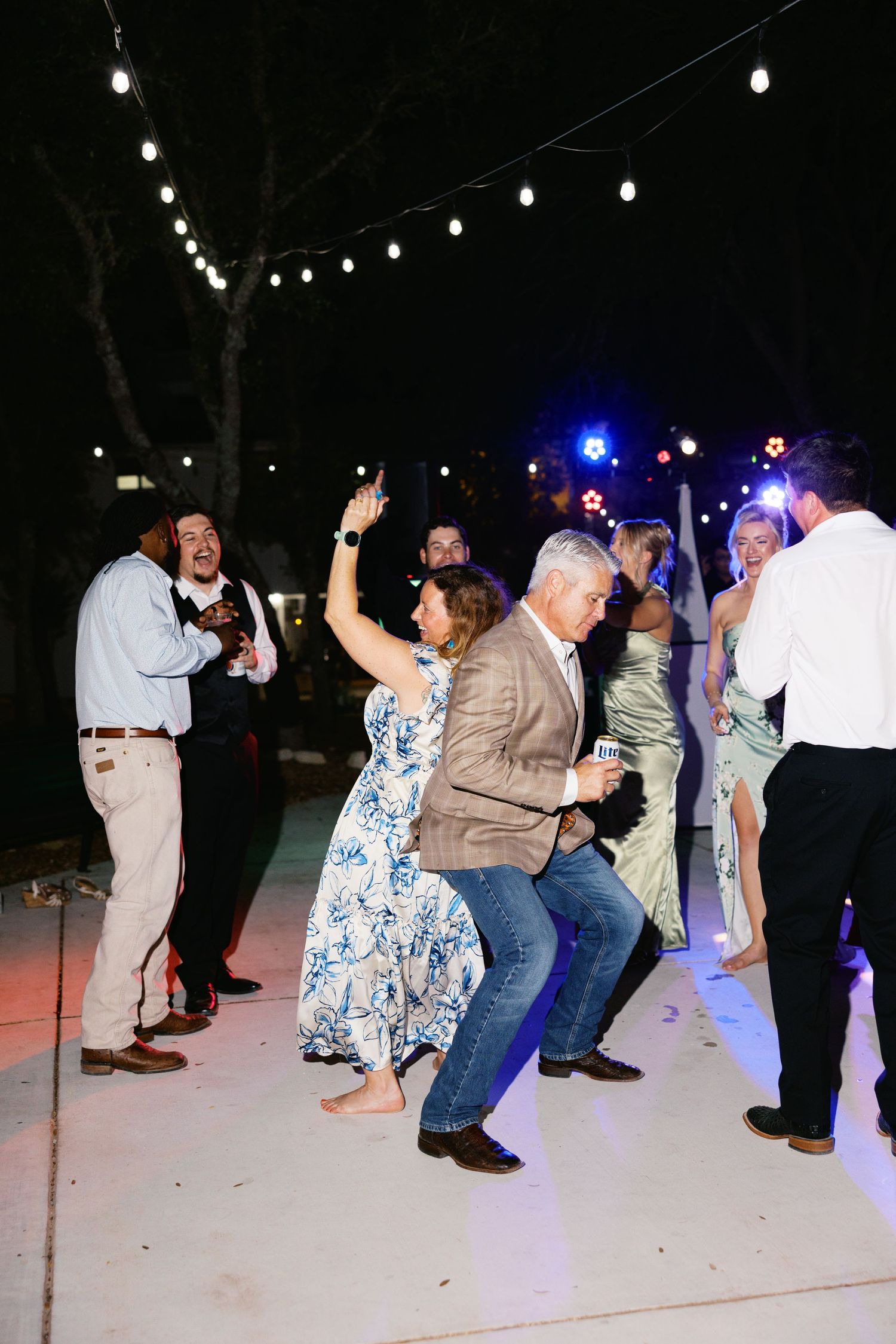Wedding guests dance energetically under string lights at an outdoor nighttime wedding reception party.