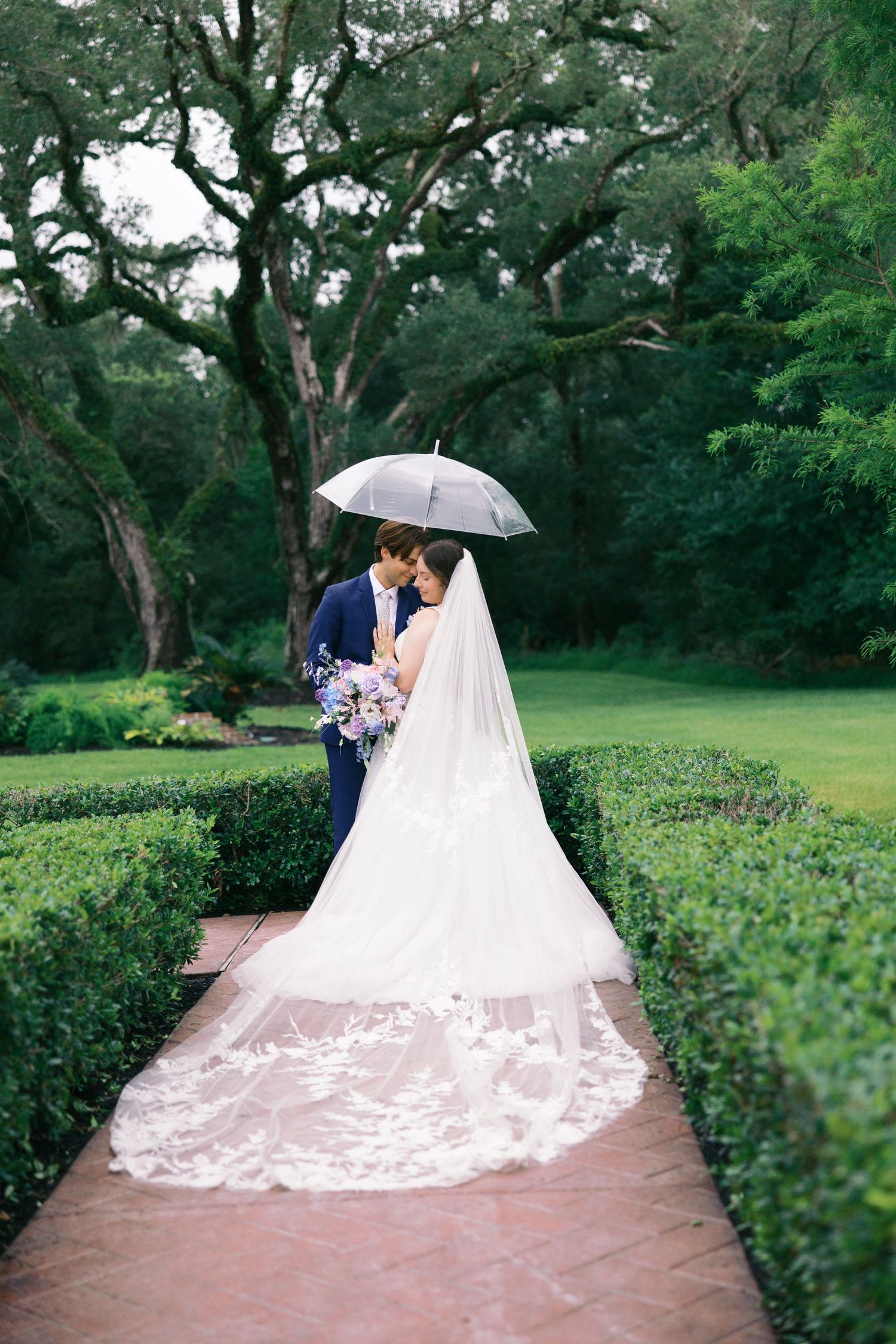 Newlyweds sharing an intimate moment under an umbrella on a garden path with a flowing lace train.