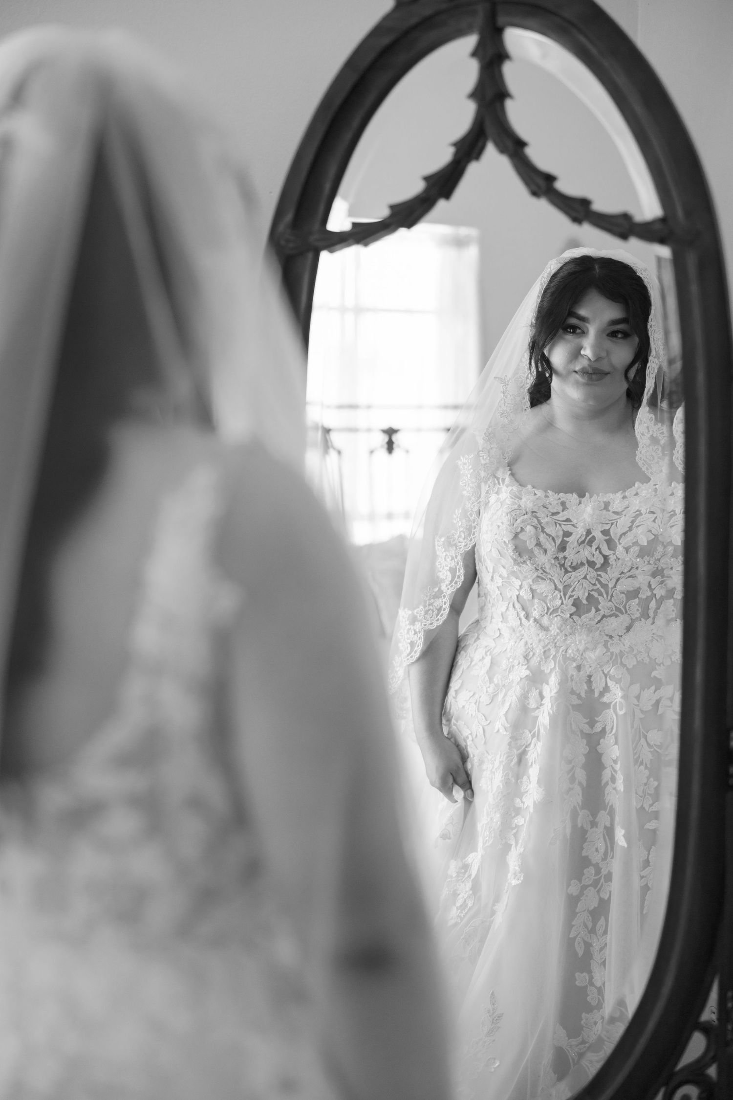 A bride admires her reflection in an ornate vintage mirror while wearing a lace wedding gown.