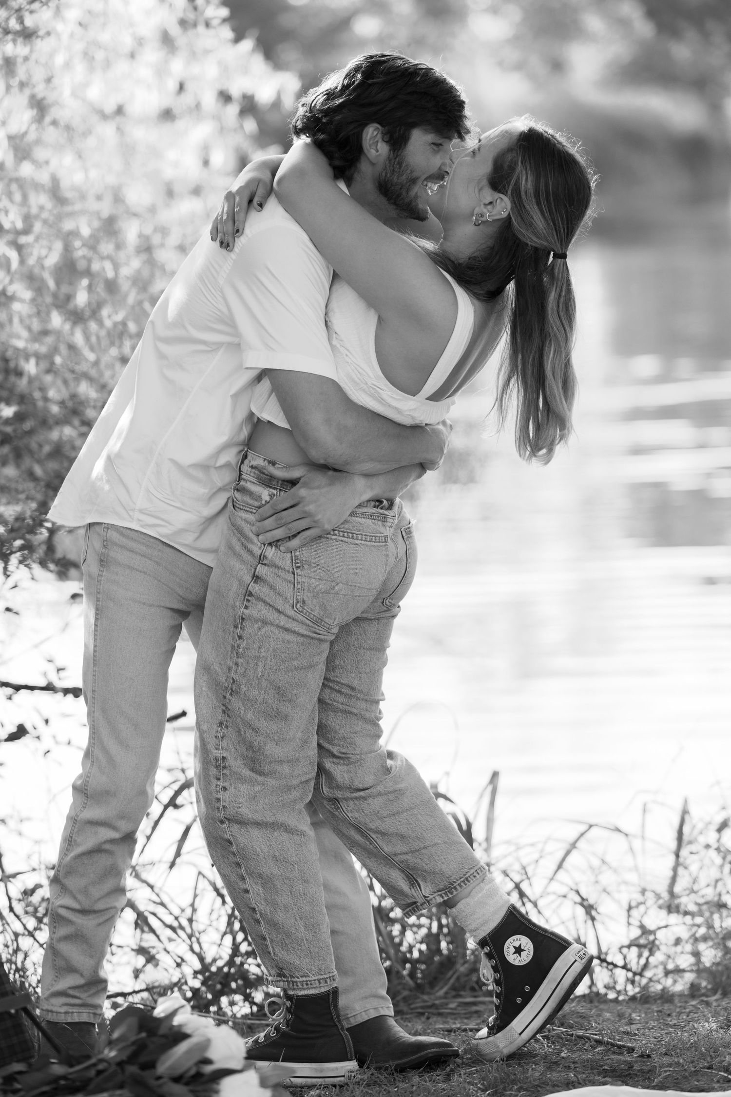 A black and white portrait of a couple in casual wear sharing an intimate moment by the water.