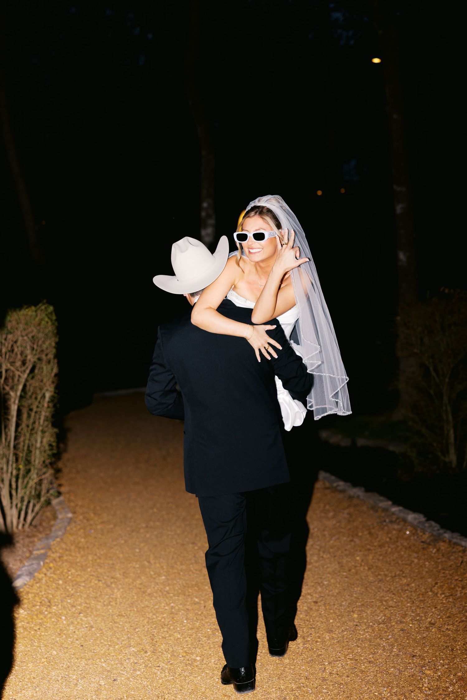 Bride in white veil and sunglasses embraces groom in cowboy hat on a lit pathway during nighttime reception.