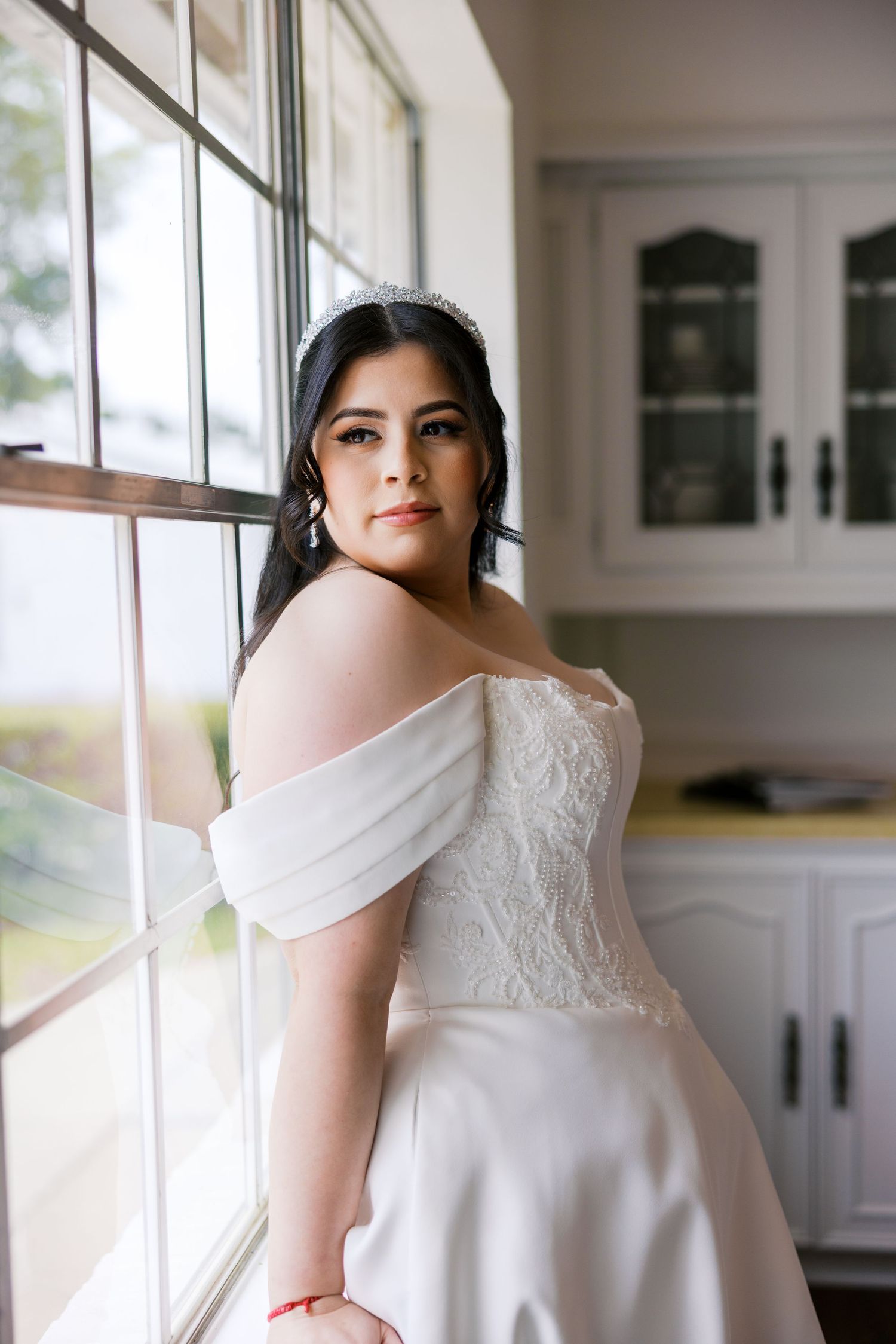 Bride in off-shoulder white lace wedding gown with tiara poses by window in elegant indoor setting.