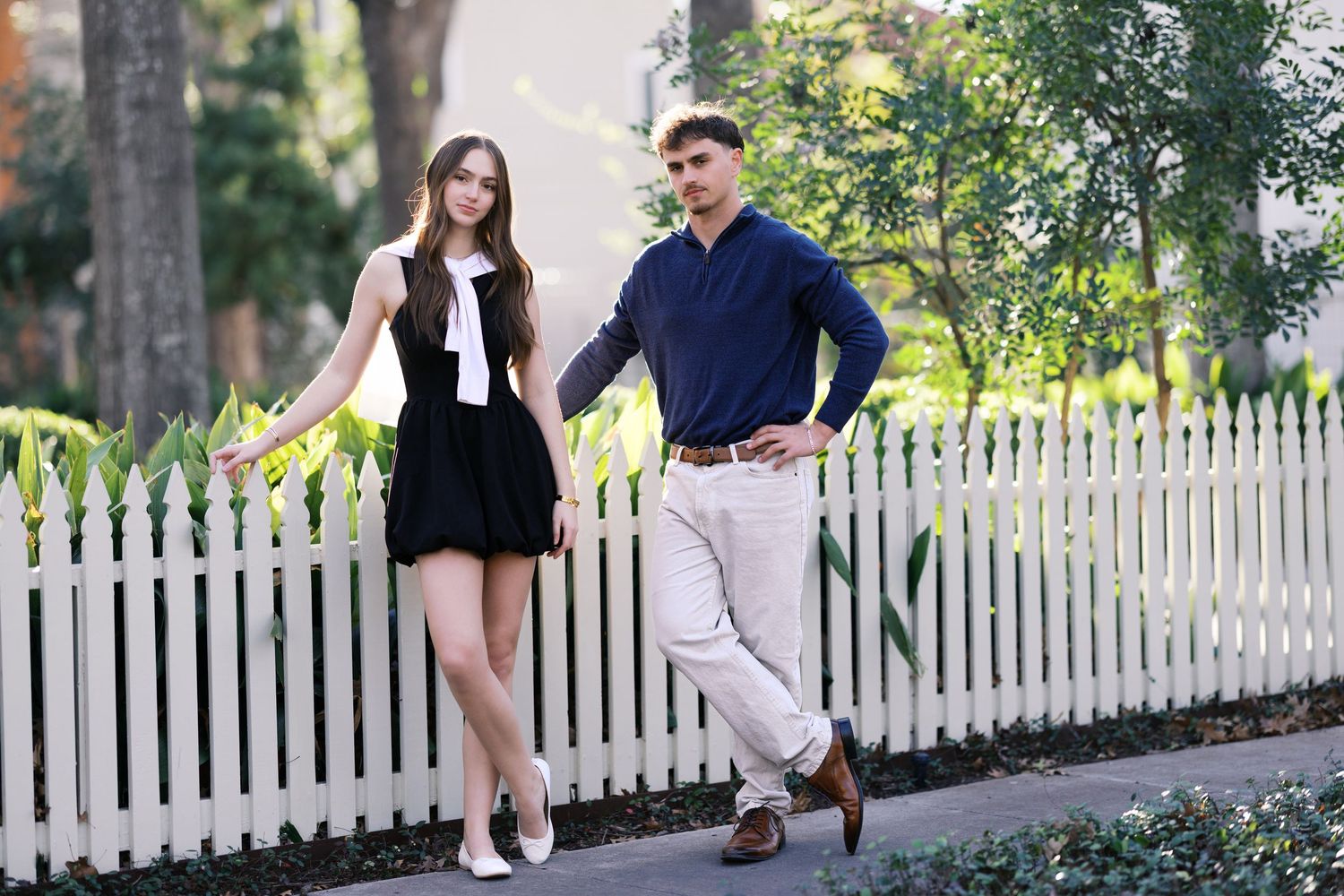 A young couple poses by a white picket fence outdoors, woman in black dress, man in navy sweater and khakis.