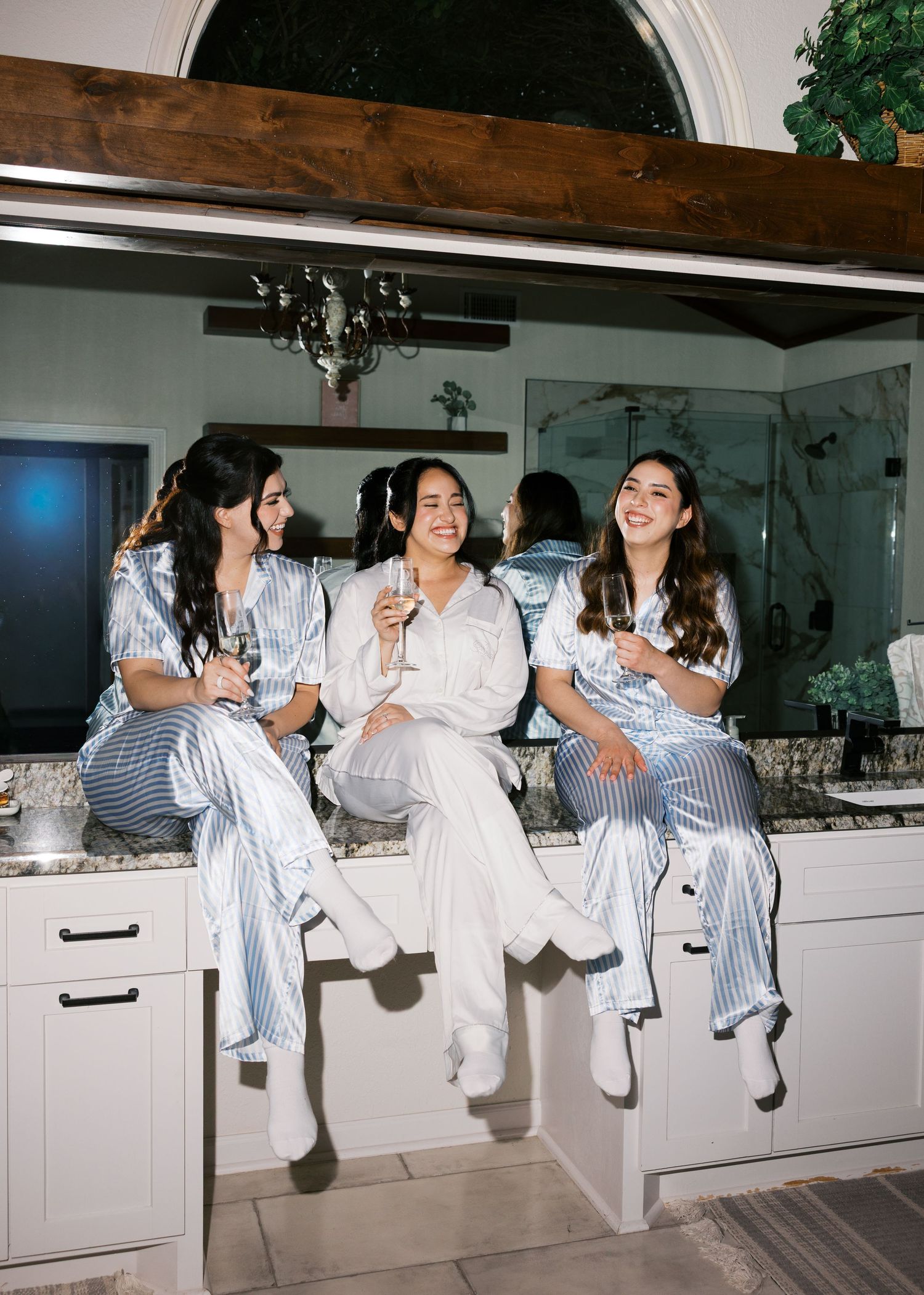 Bride and bridesmaids in matching pajamas sitting on bathroom counter celebrating with champagne glasses.