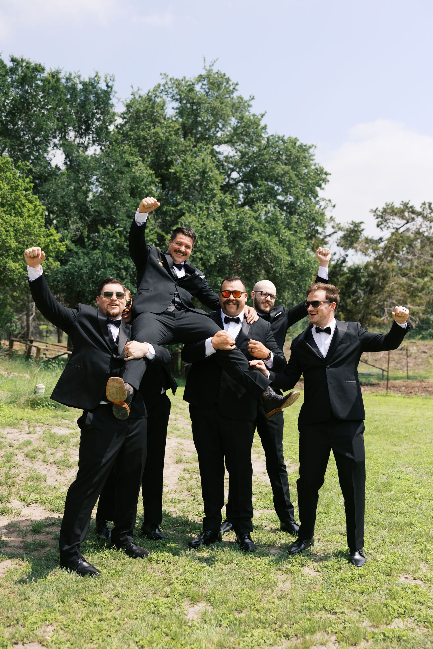Groomsmen in black tuxedos lifting groom outdoors, celebrating with raised fists at a wedding.