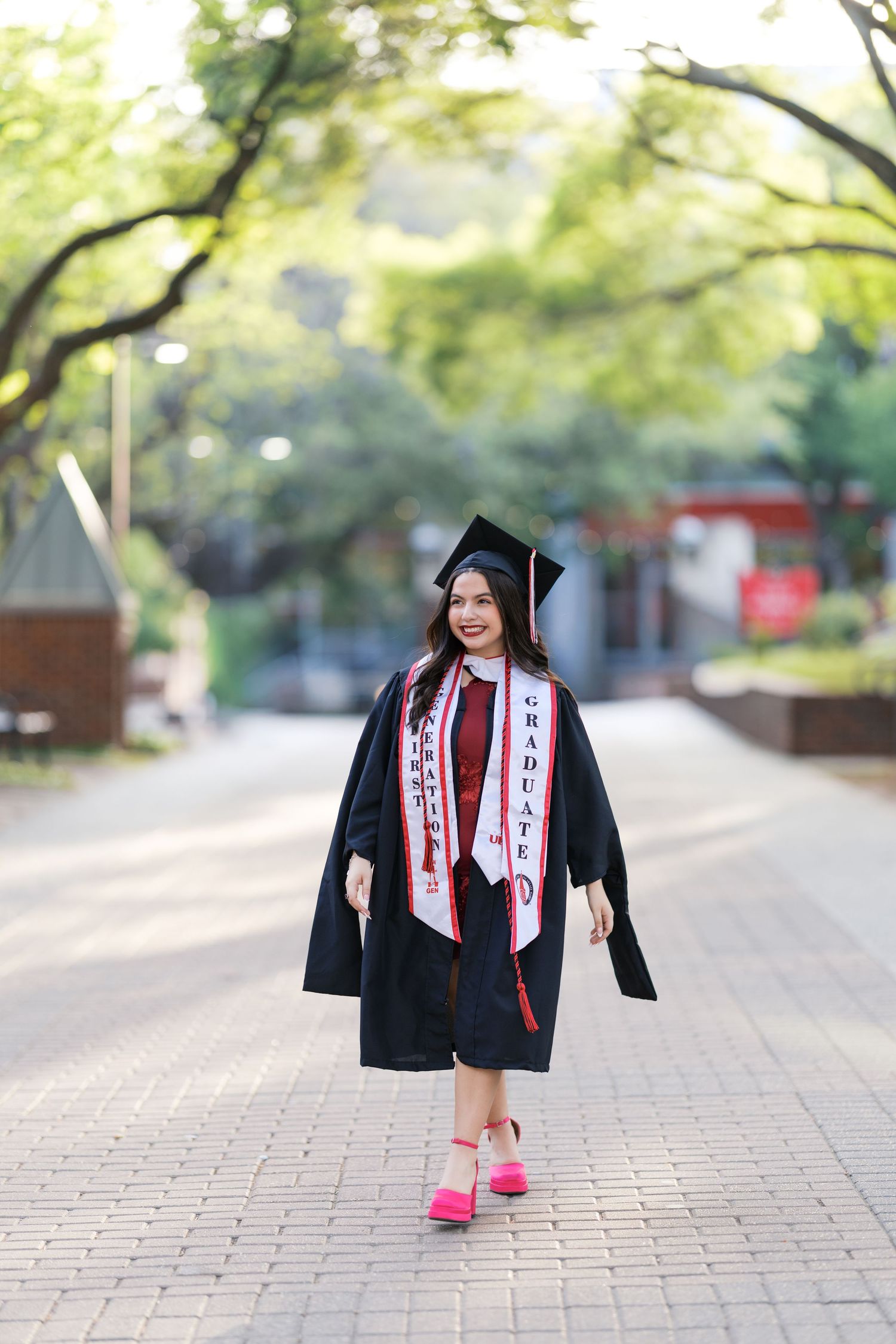 Smiling graduate in black cap and gown with honor stoles walking on a tree-lined campus path.