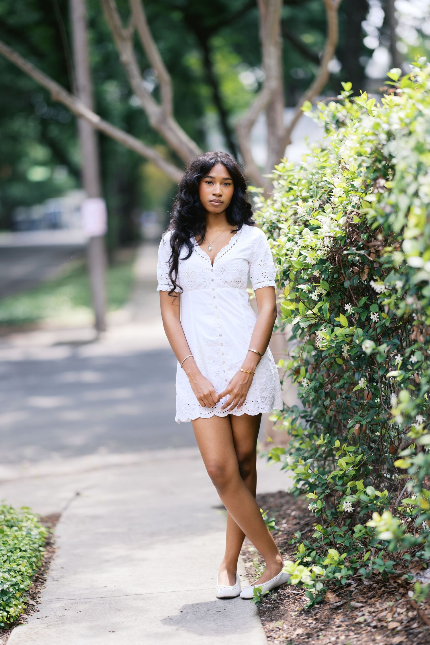 Young woman in white eyelet mini dress standing on a sidewalk beside lush green bushes.