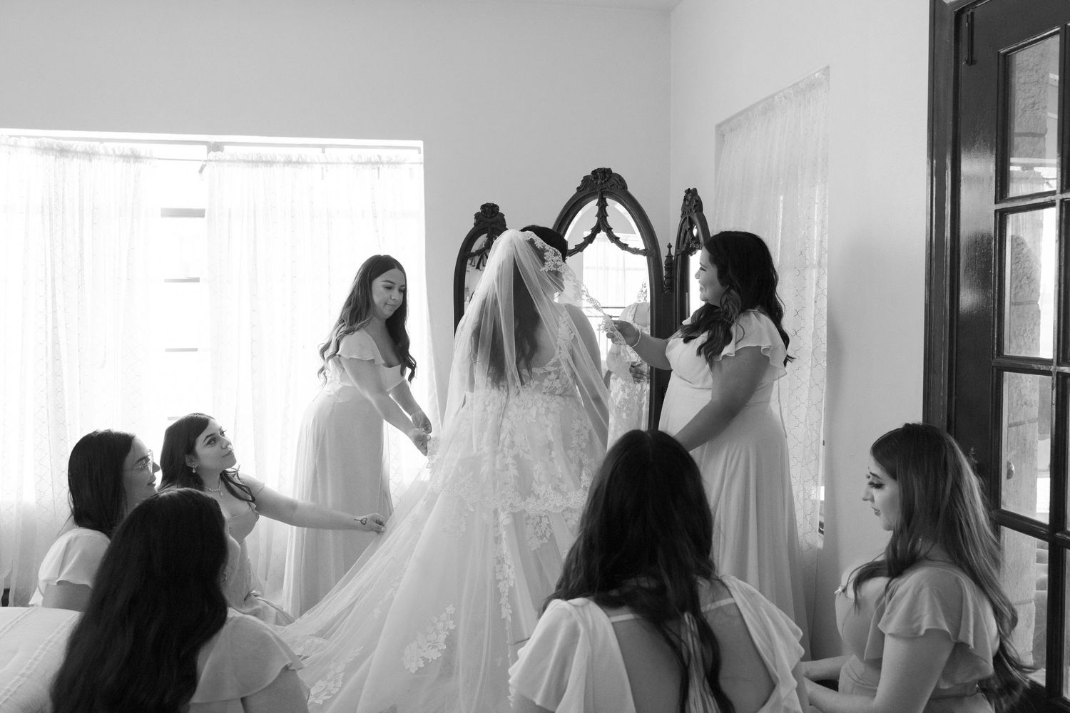 Bridesmaids in white robes help the bride with her veil during wedding preparations.