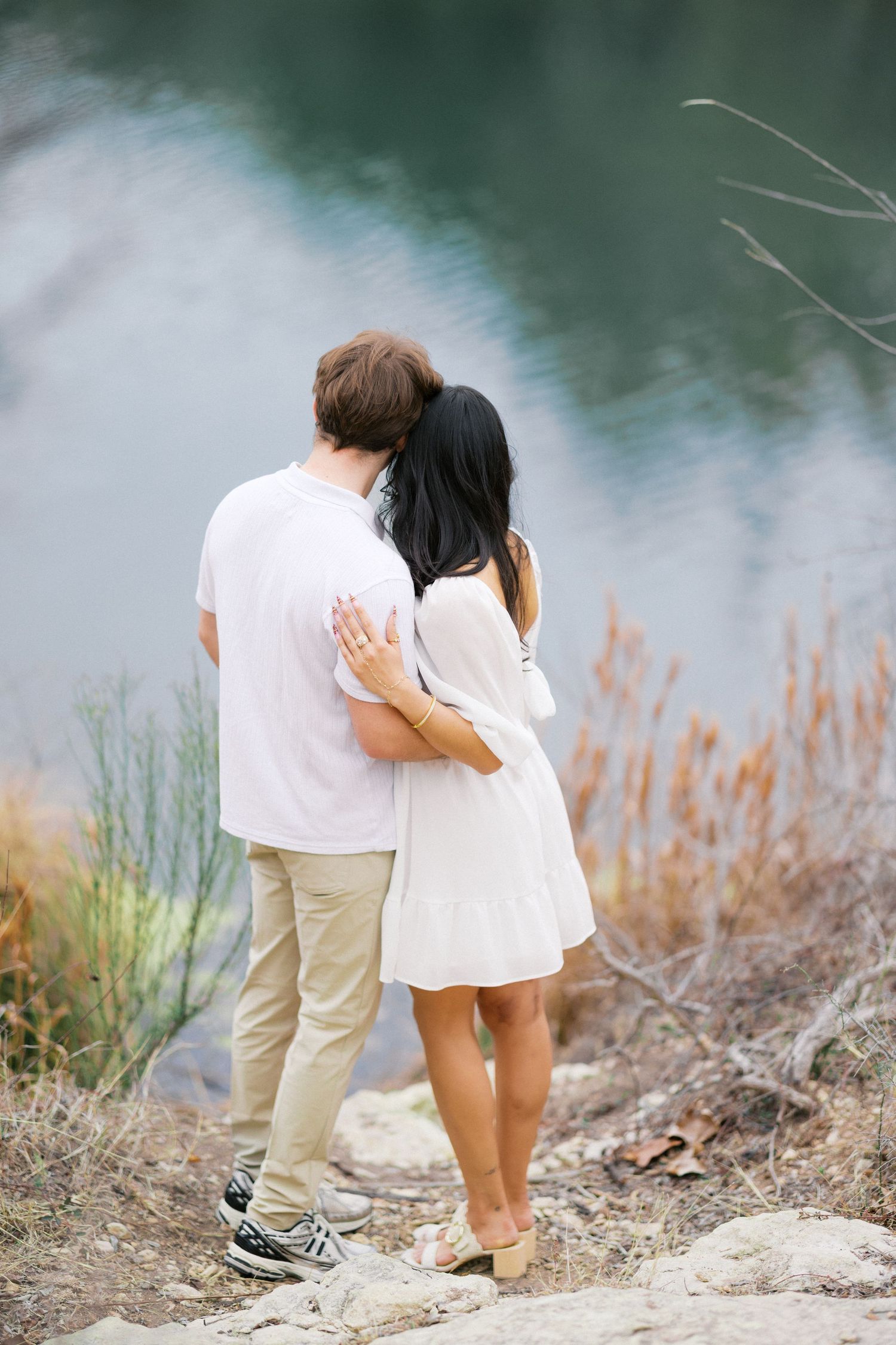 Couple walking together along rocky shoreline with calm water and natural vegetation in background.