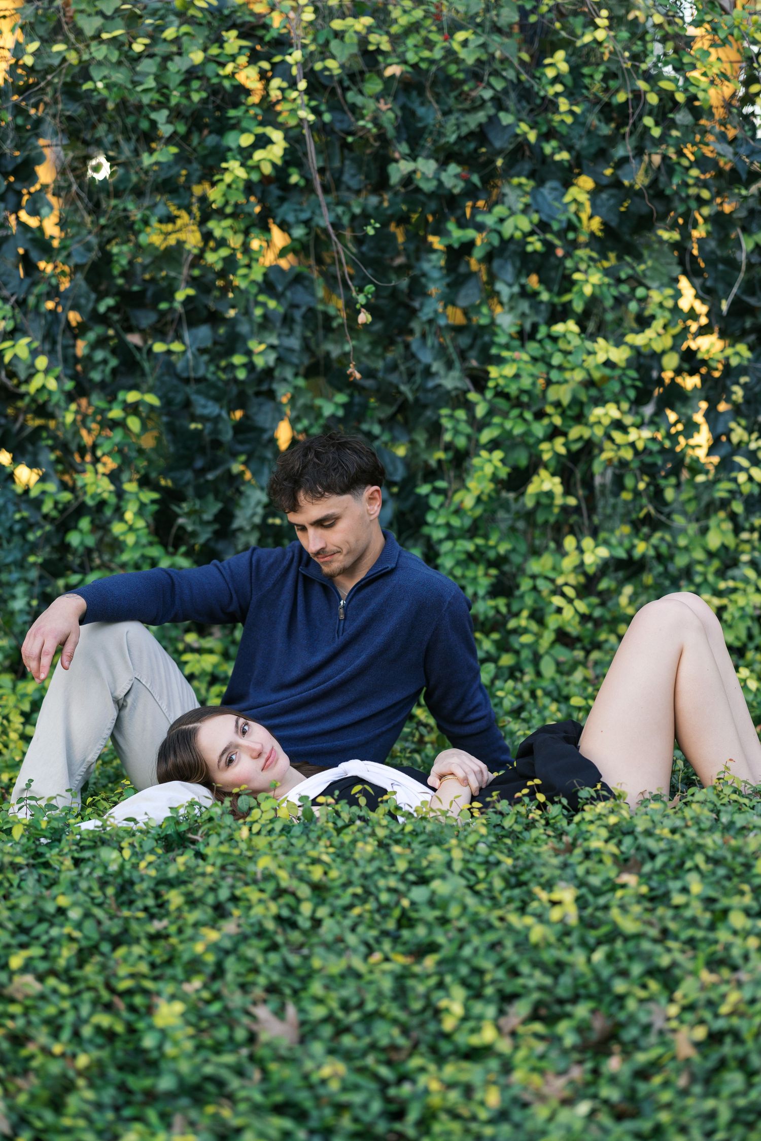 Romantic couple relaxing together on lush green ivy hedge in sunlit outdoor garden setting.