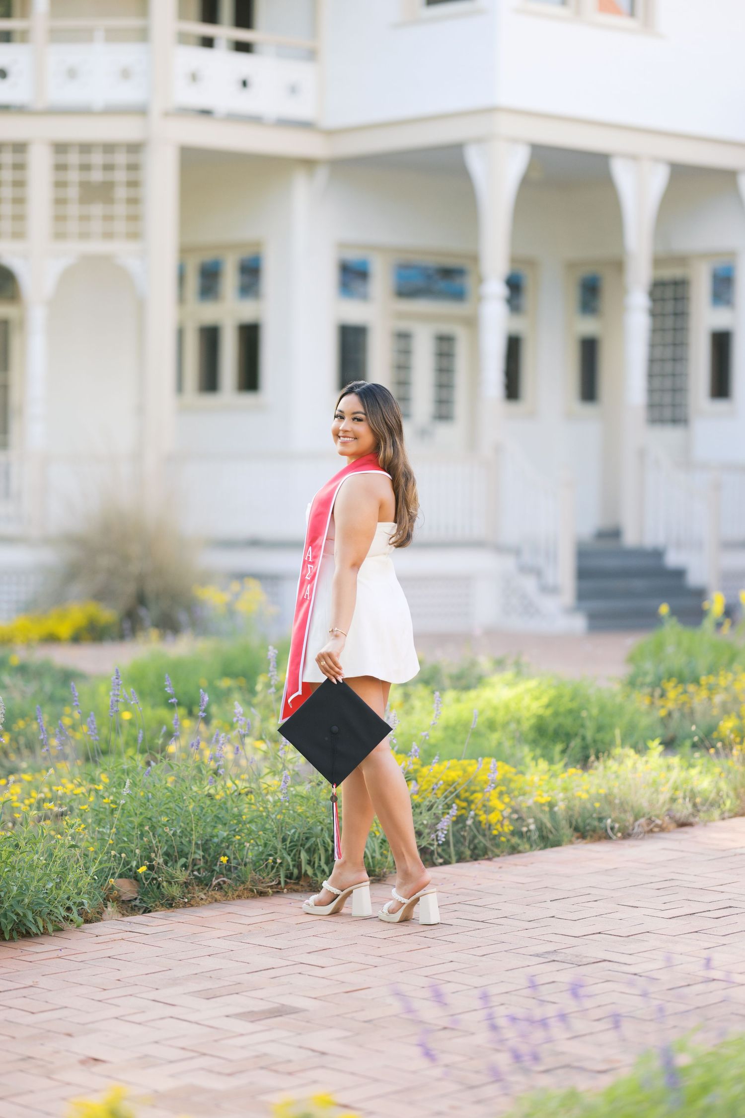 Young woman in white dress and red graduation sash holding cap, posing outdoors near a white building.