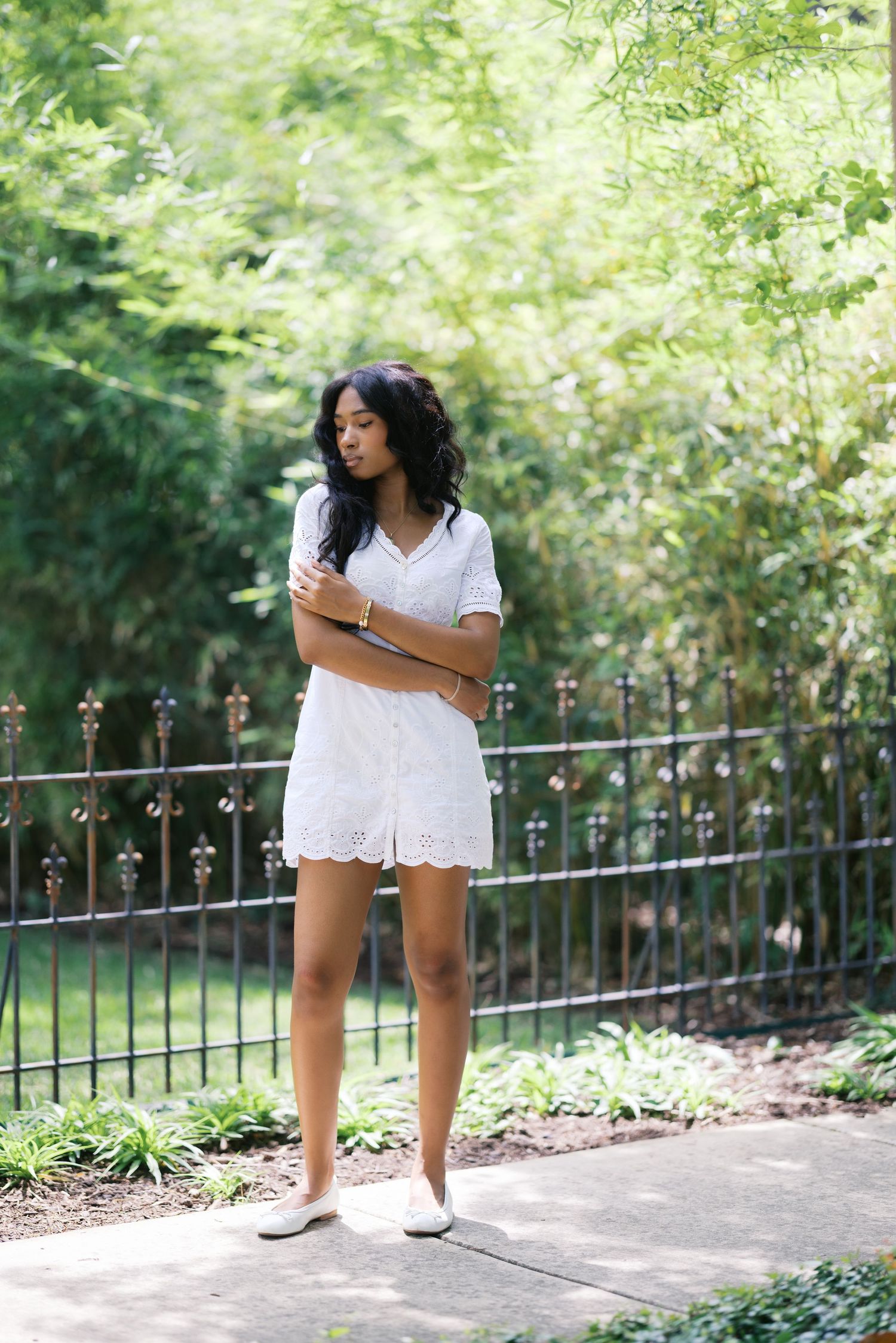 Young Black woman in white eyelet dress standing on path near black iron fence surrounded by greenery.