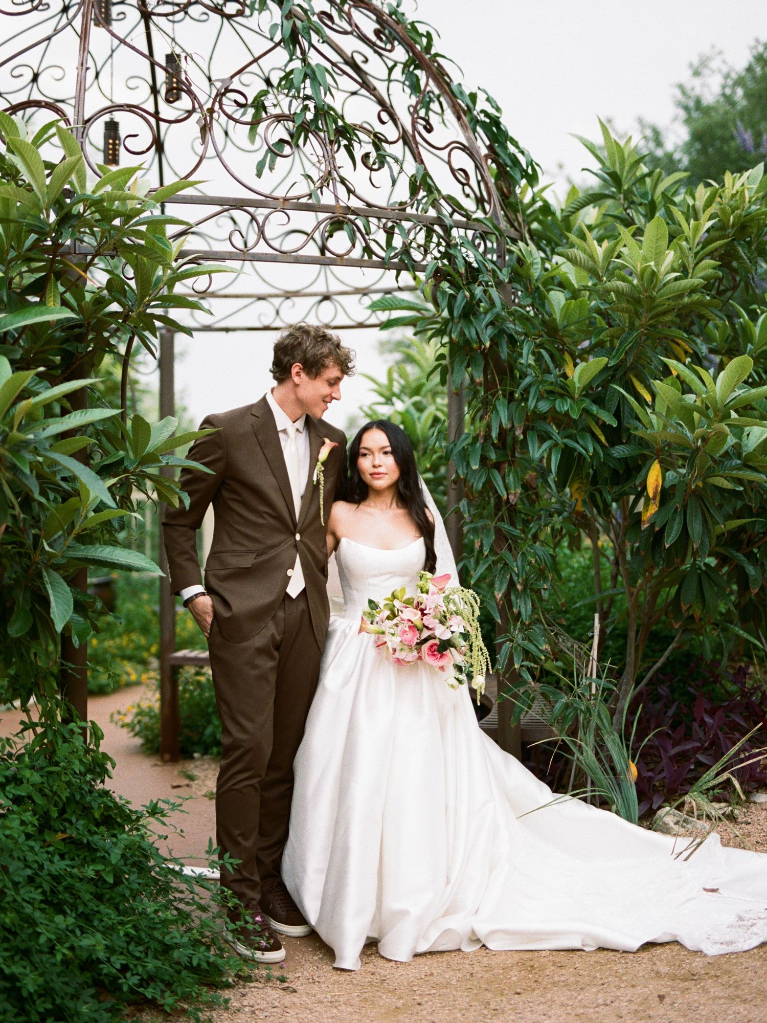 Bride and groom pose under ornate iron garden arbor, bride in white gown holding pink bouquet, groom in brown suit.