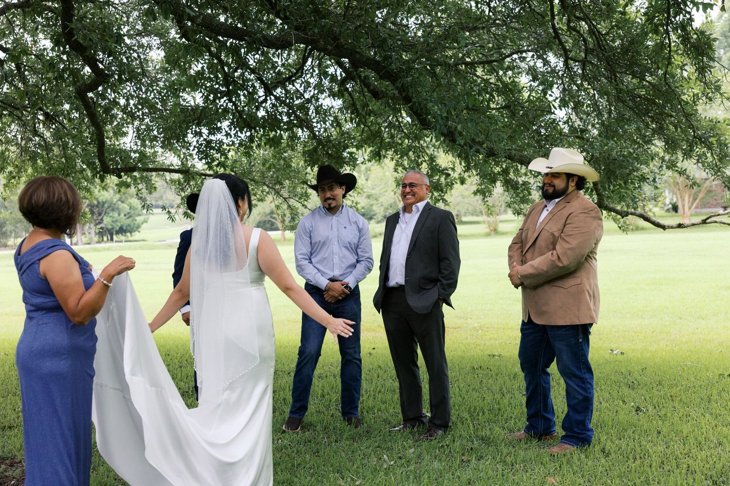 A bride in a white gown and veil meets guests under a large tree at an outdoor wedding ceremony on a green lawn.