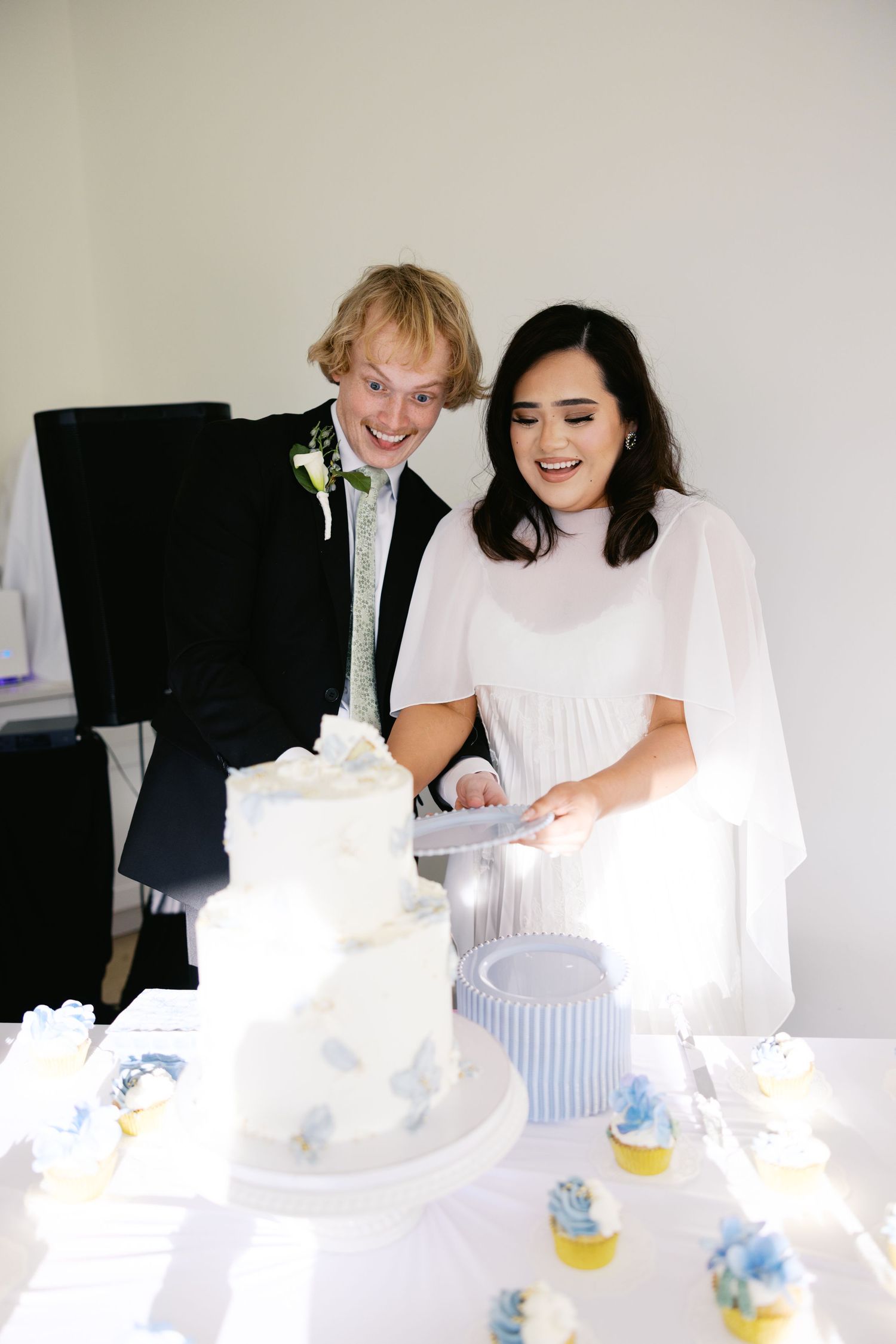 Smiling bride and groom cutting white tiered wedding cake at reception table with cupcakes.