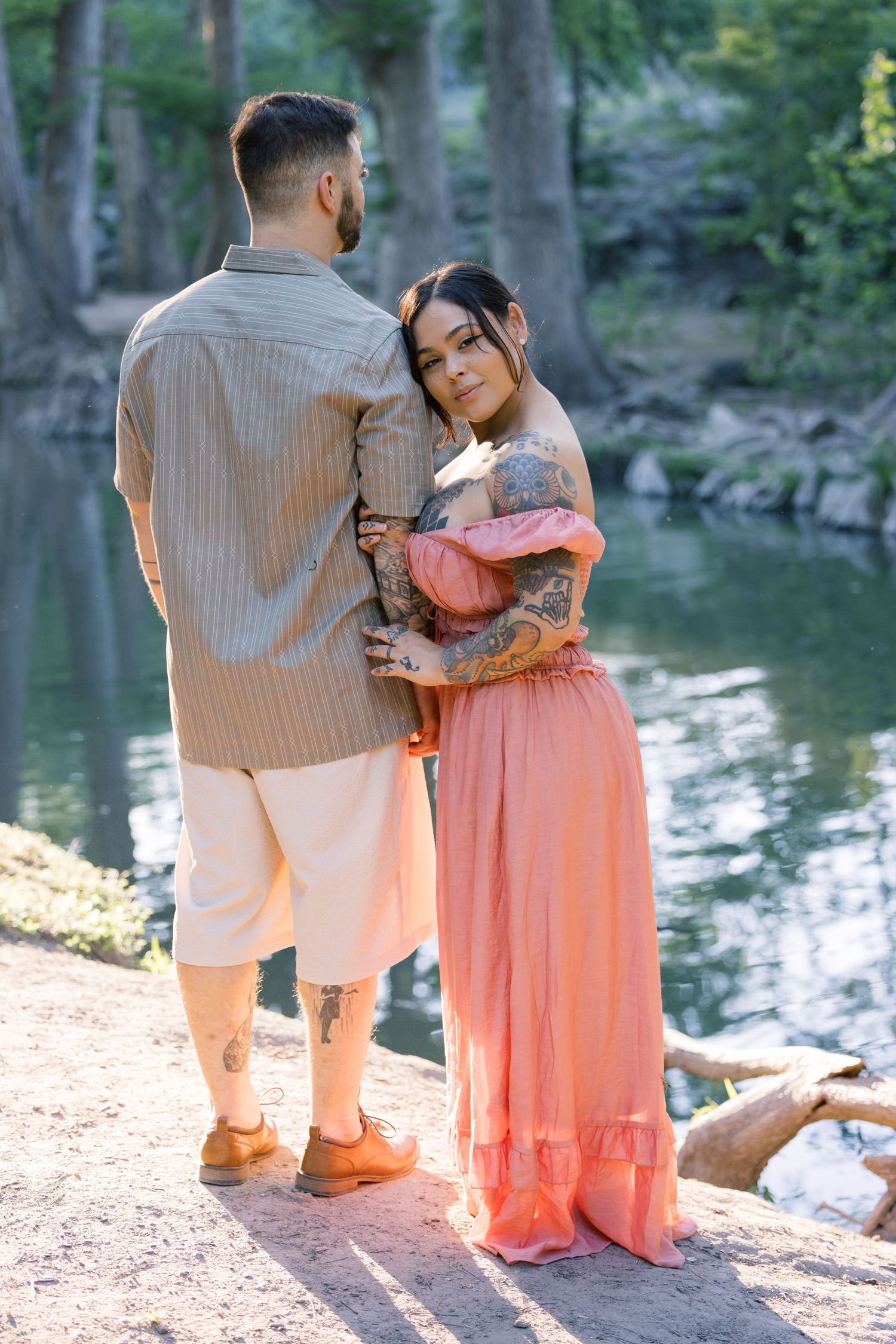 Two people embrace while standing on a rocky shore beside a tranquil lake surrounded by trees.