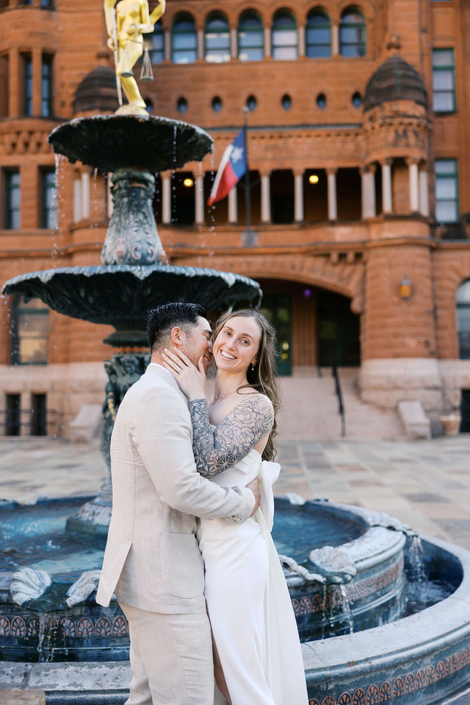 Couple embracing by ornate fountain outside historic red brick building.