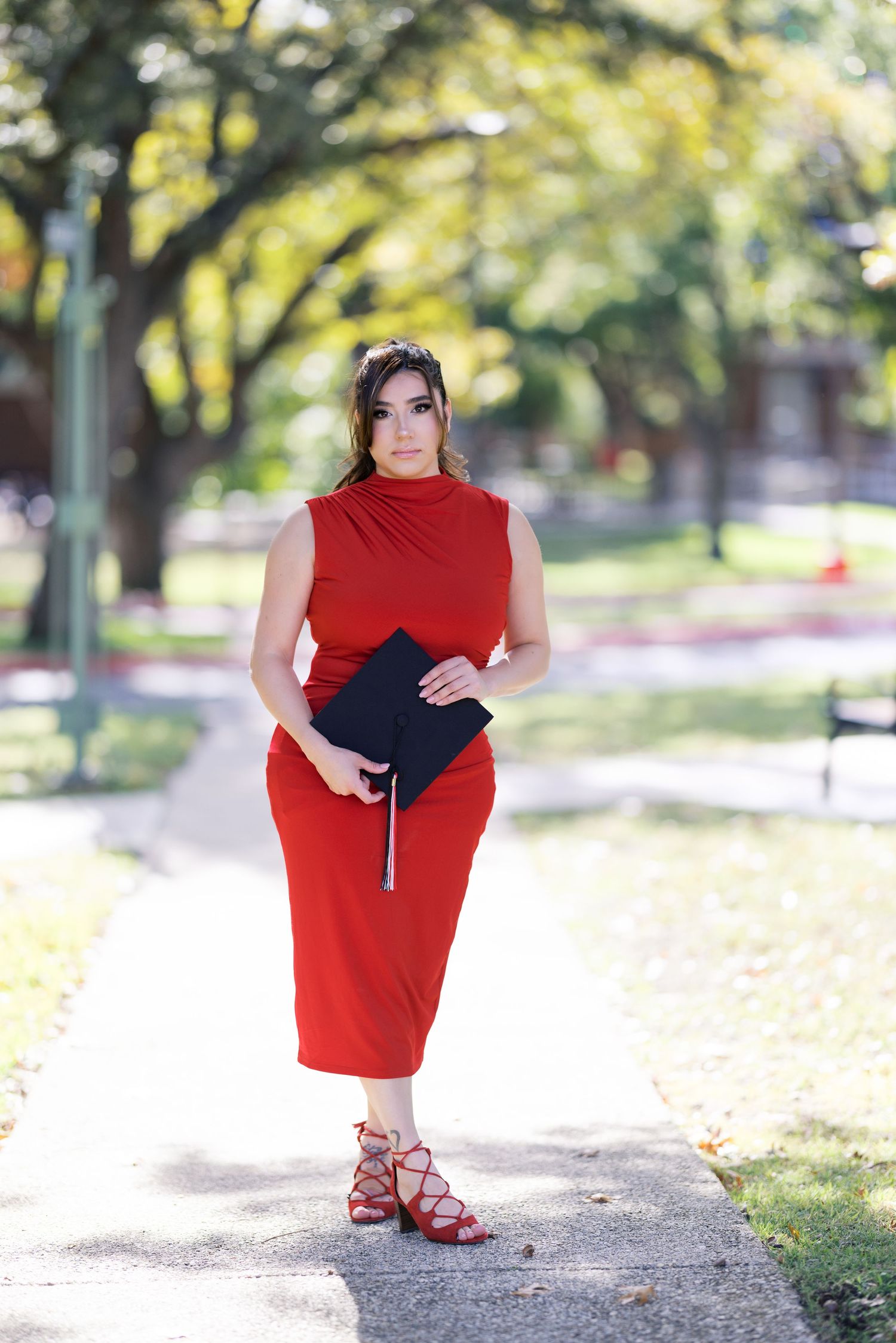 Graduate in red dress holding diploma while walking through park setting.