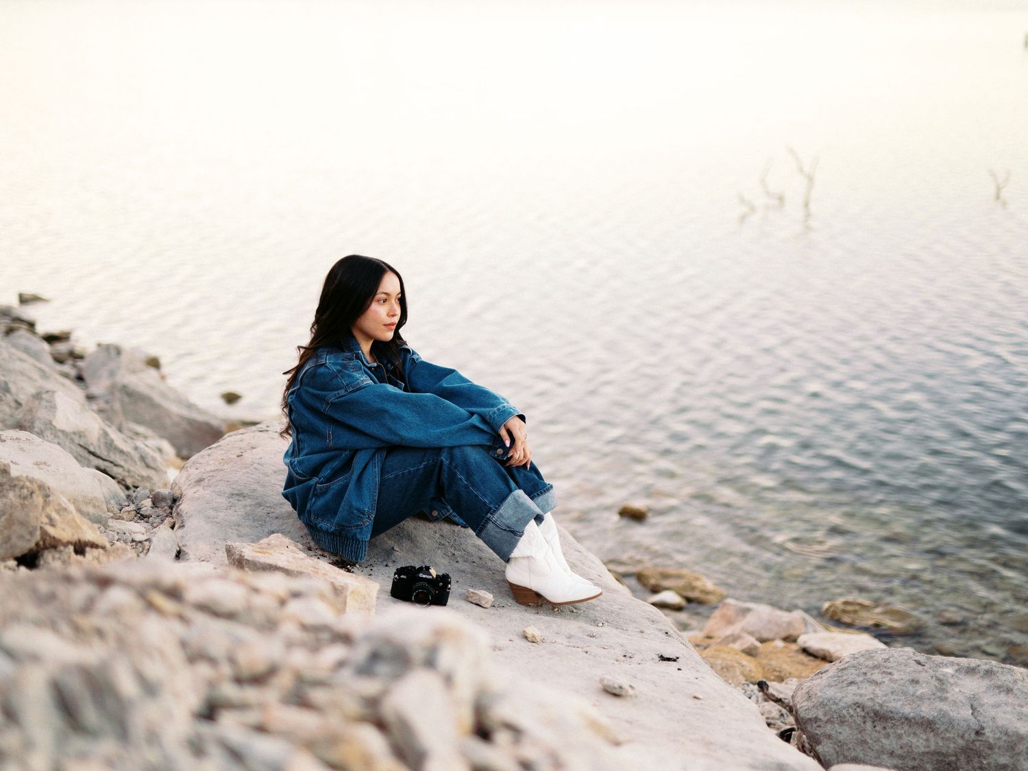 Young woman in denim jacket sitting on rocky shore gazing at calm water during golden hour.