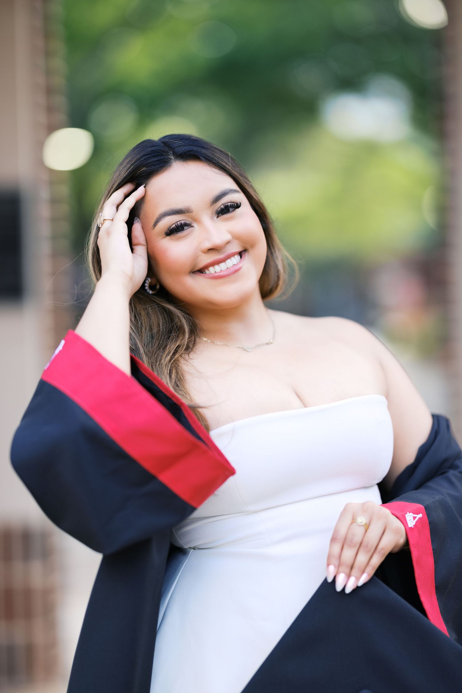 Smiling graduate in white dress and black graduation gown poses outdoors for senior portrait photo.