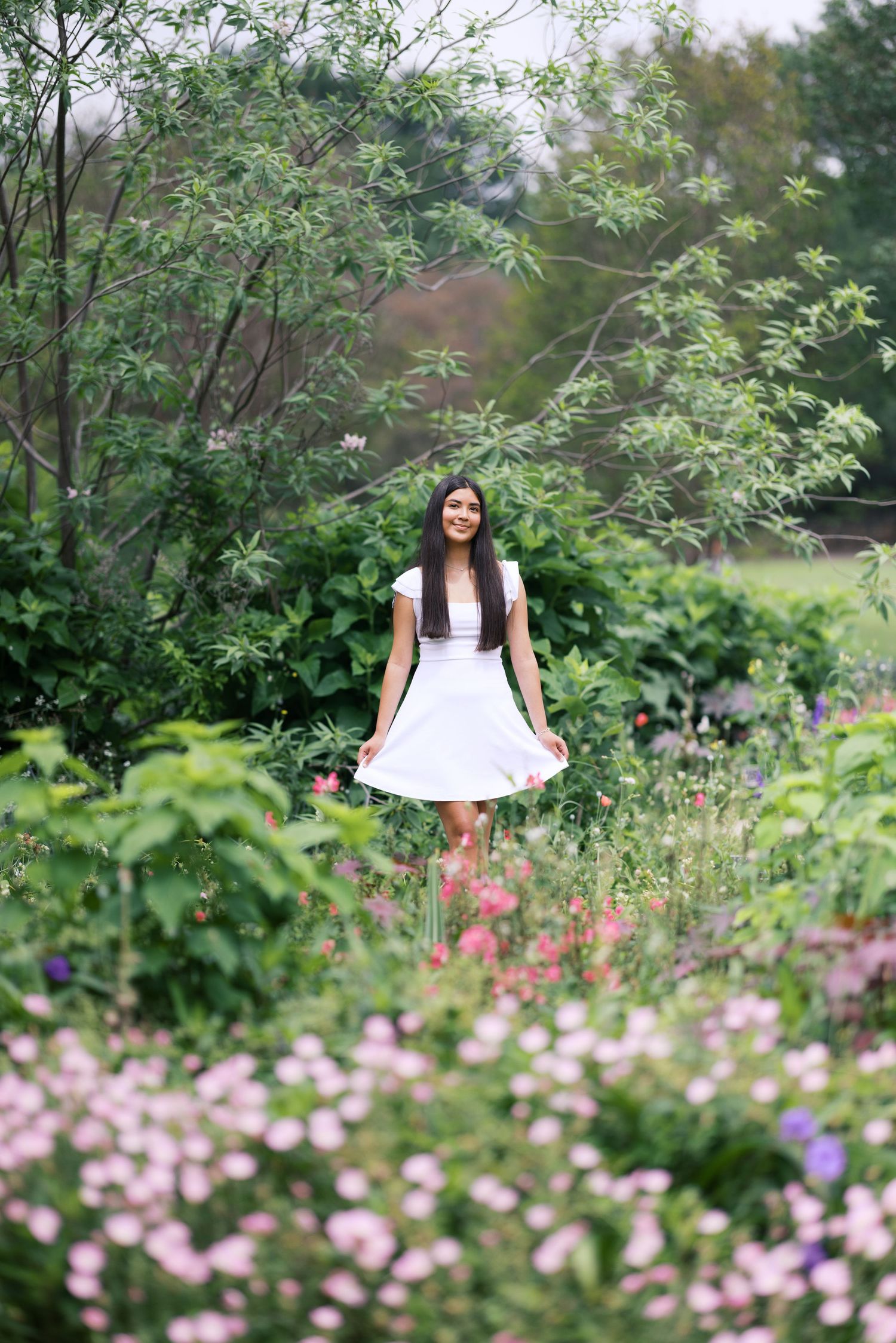 Young woman in white dress standing among colorful blooming flowers in a lush green garden.