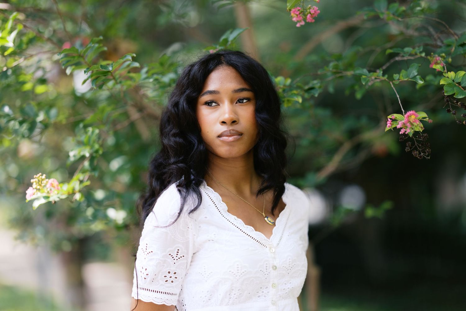 Young Black woman with long wavy hair wearing a white lace dress standing outdoors among green trees and pink flowers.