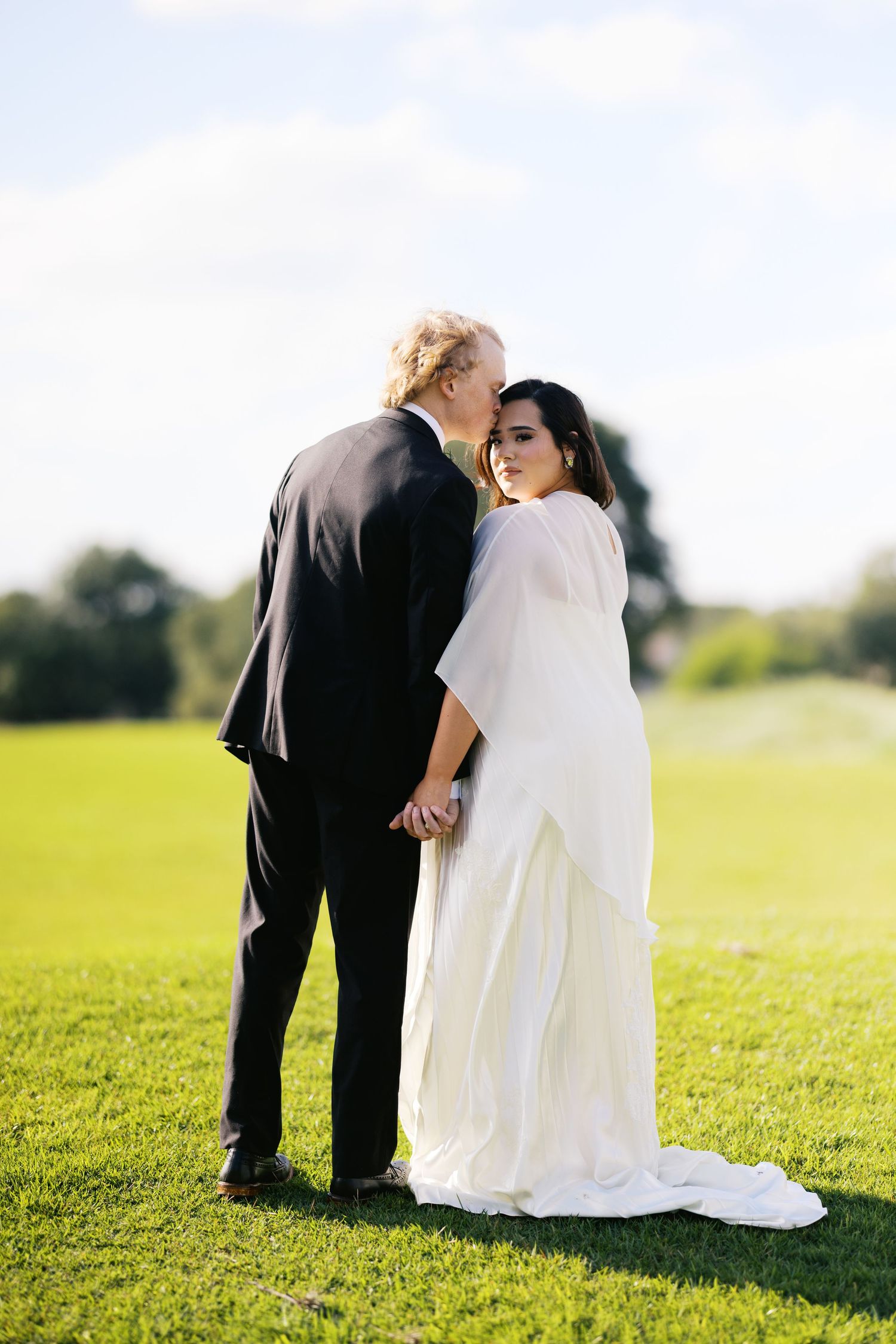 A bride in a white gown and groom in a black suit hold hands on a green lawn, sharing a tender moment.