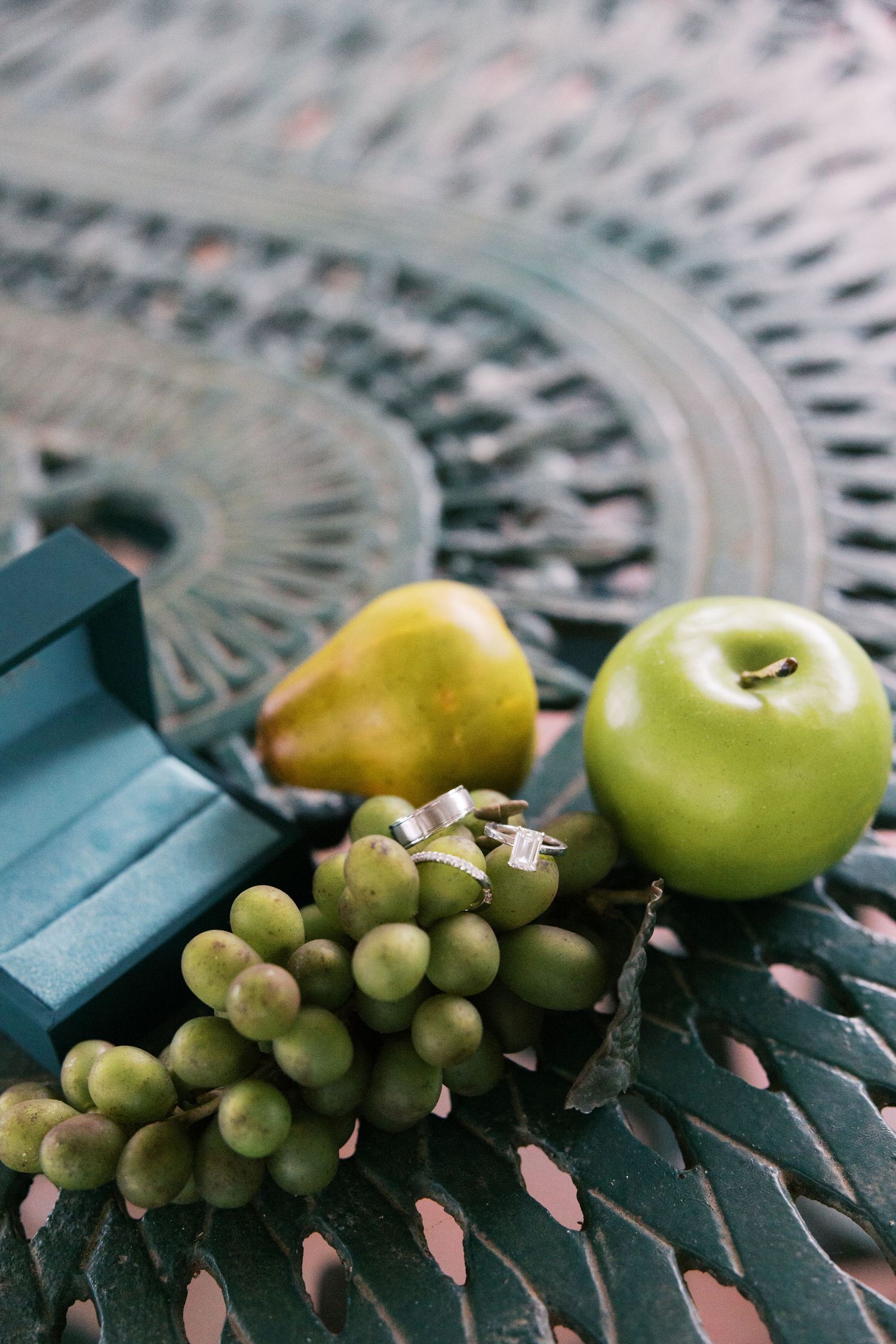 Wedding rings displayed beside green grapes, pear, and apple on ornate metal table.