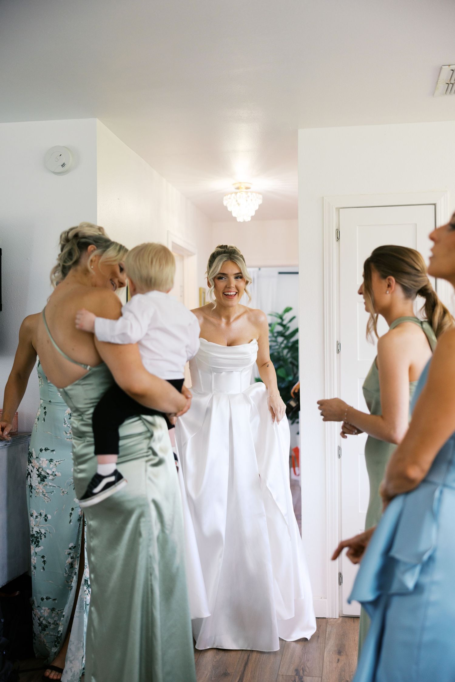 Bride in white strapless gown smiles at bridesmaids in sage green dresses during wedding preparations indoors.