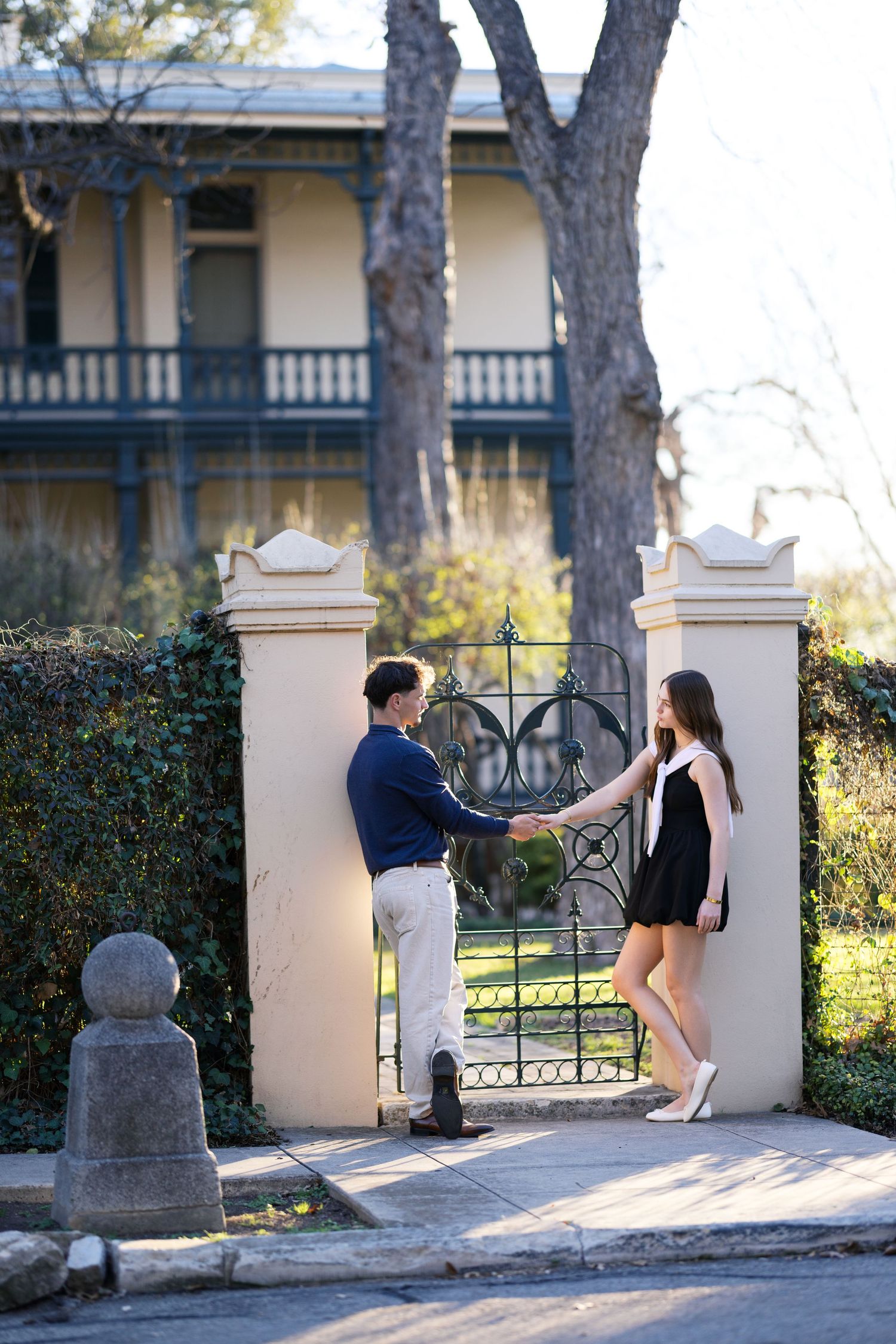 Young couple holding hands at decorative iron gate outside charming historic home with balcony.