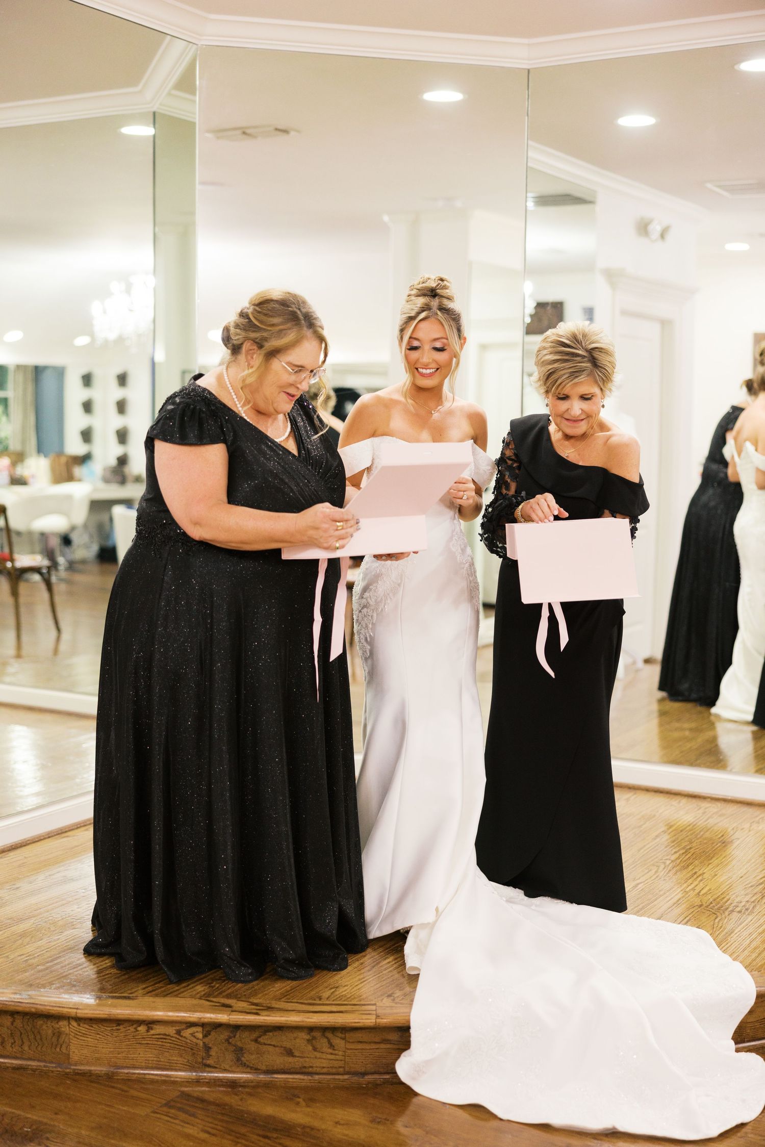 Bride in white wedding dress opening pink gift box with two women in black formal gowns.