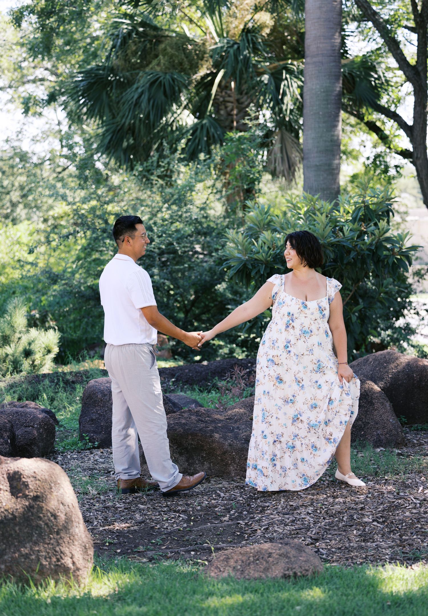 A couple holds hands while walking through a garden path surrounded by rocks and palm trees.