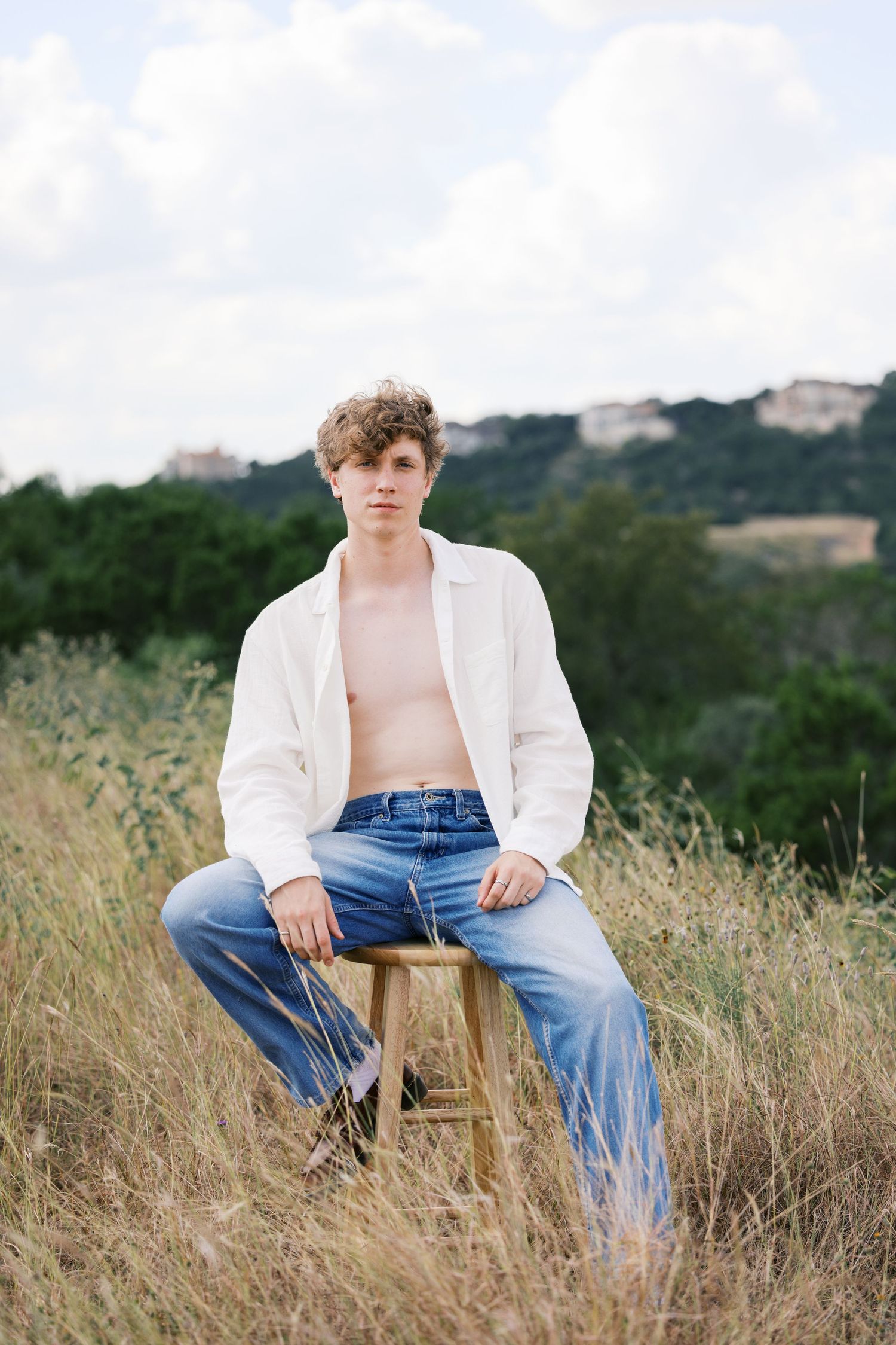 Artistic portrait on wooden stool in field wearing unbuttoned white shirt and jeans.