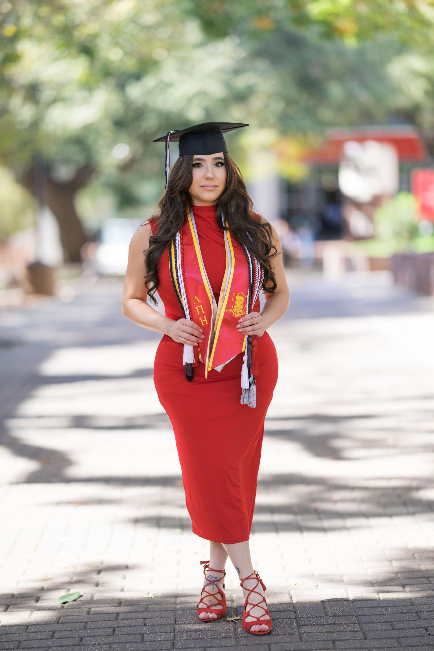 Graduate in red dress and graduation cap poses outdoors on brick walkway.