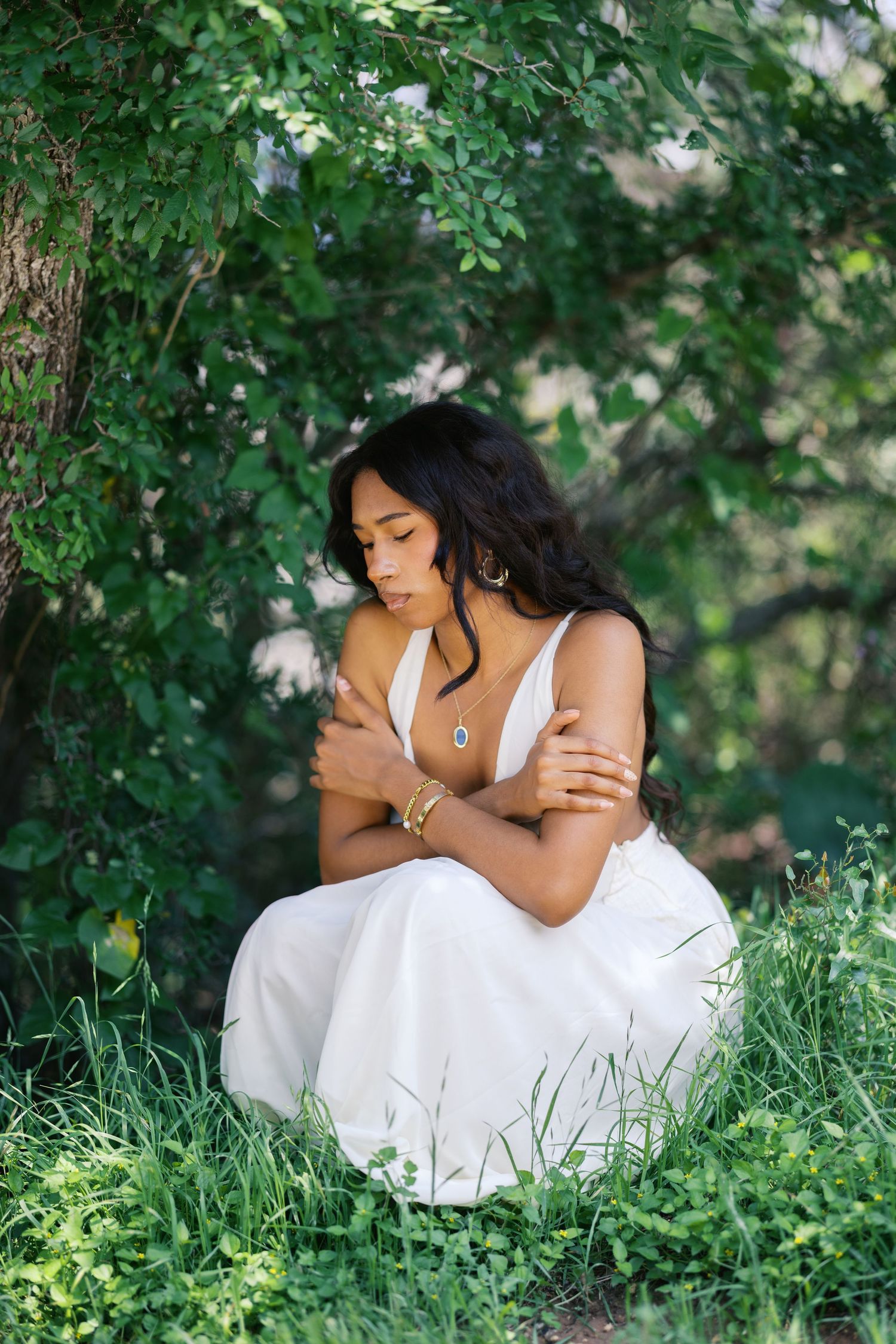Young woman in white dress sitting peacefully in lush green grass beneath a leafy tree.